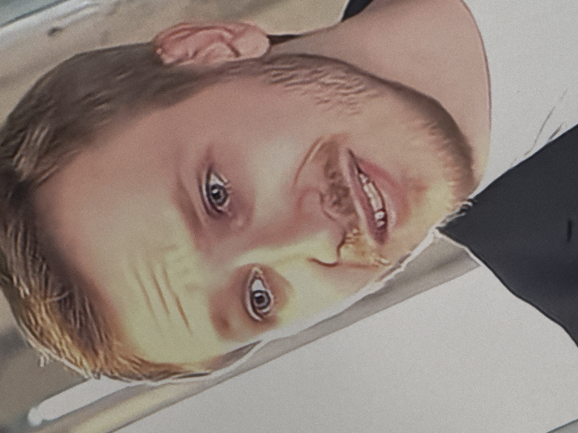 The image is a close-up, slightly blurred, medium-shot portrait of a young Caucasian male with light brown hair and blue eyes, looking slightly off-camera. He has a light beard and a faint smile, revealing some teeth. He is wearing a dark jacket over a white t-shirt. The background is indistinct and appears to be an indoor setting with a light-colored wall and a vertical element that could be a pillar or a light fixture. There is no identifiable information suggesting a location in Saratov, Russia.