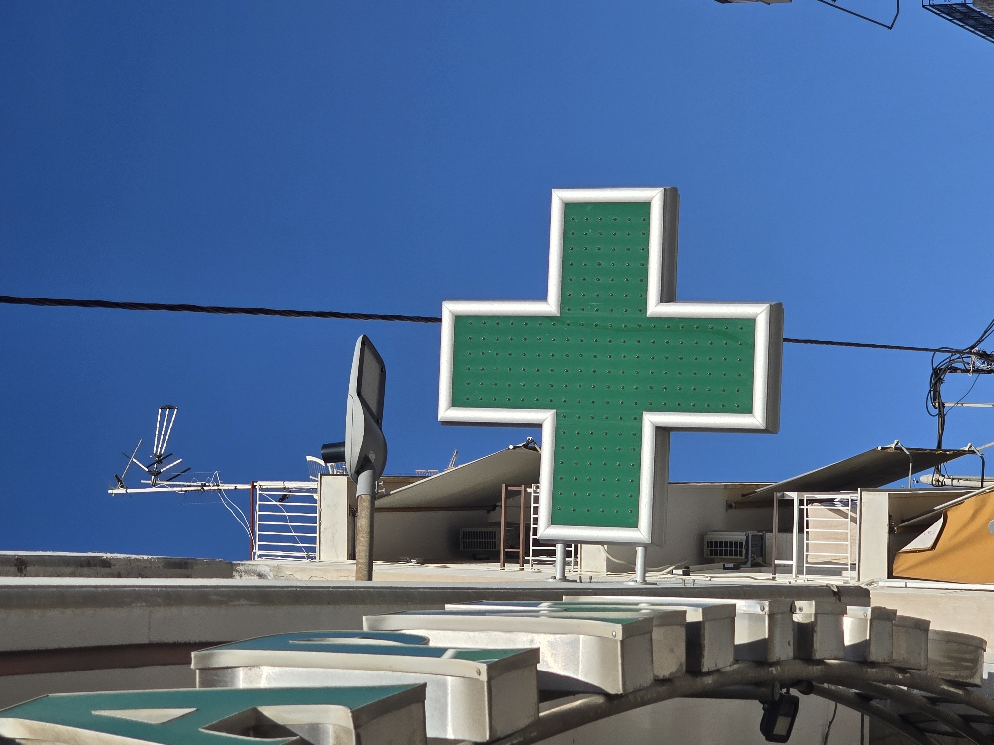 A green cross pharmacy sign is mounted on the side of a building in Athens, Greece. The sign is illuminated and has a white border. The building appears to be made of concrete and has a balcony with a white railing. There is also an air conditioning unit visible on the balcony. A television antenna is mounted on the roof of the building. The sky is clear and blue. The image is taken from a low angle, looking up at the sign and building.