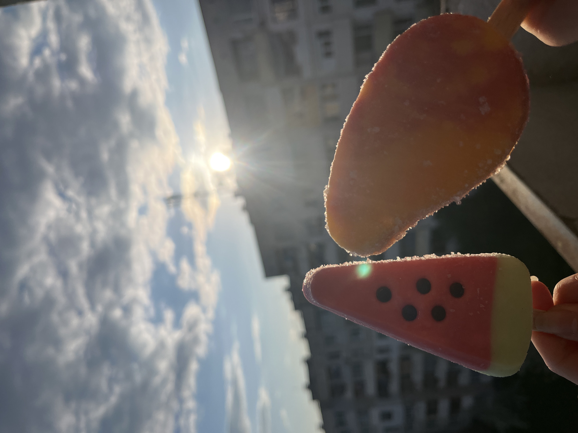 The photograph is rotated 90 degrees clockwise, presenting a vibrant outdoor scene with two ice pops held against a bright sky and a blurry urban backdrop. In this orientation, the sun is a brilliant disc positioned in the top right corner, emitting strong rays and lens flare that illuminate the subjects.
In the foreground, partial views of hands hold two distinct popsicles. The upper popsicle is elongated, displaying shades of orange and yellow, possibly mango-flavored, and is visibly covered in fine ice crystals, suggesting its cold temperature and beginning to melt slightly. Below it, a second popsicle is shaped like a triangular watermelon slice, with a red top portion featuring several black "seeds" and a light green/yellow rind at its base. This popsicle also shows a frosty surface.
The background on the left is dominated by a clear blue sky interspersed with fluffy white clouds. On the right, a tall, multi-story building with many windows is rendered in soft focus, indicating an urban environment, likely in Sofia, Bulgaria. The image captures a moment of summer enjoyment and refreshment on a sunny day. No text is visible.