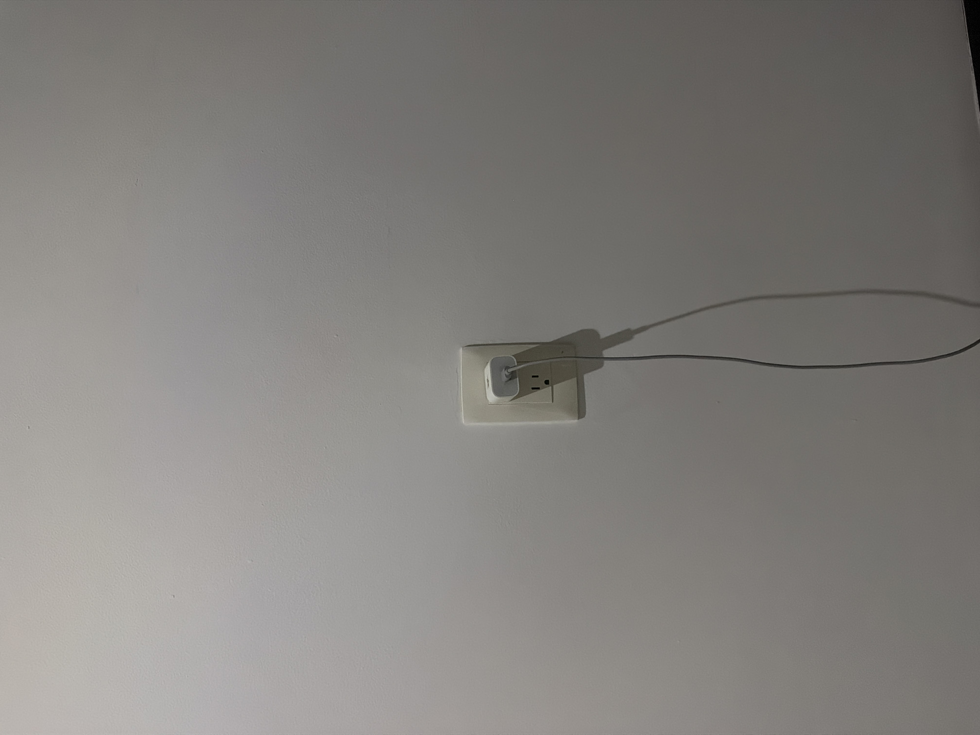 A white wall contains an electrical outlet with a white charger plugged into it. A gray charging cable extends from the charger. The environment appears indoors. There is no verifiable information about the location being in Villahermosa, Mexico. No people, subjects, or actions are visible.