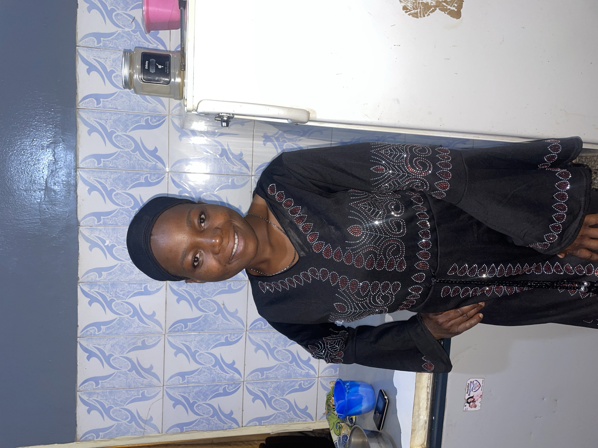 A woman stands indoors against a wall of blue and white patterned tile and a white wall. The woman is wearing a black garment with what appears to be rhinestone embellishments. She is smiling and looking at the camera. To her left, a white refrigerator is visible. On the counter below, are various objects including a blue plastic cup and a phone. A small poster or sticker is visible on the lower right of the image on the white wall. The image appears to be taken indoors in a domestic setting. There is no information to confirm this image was taken in Jos, Nigeria.