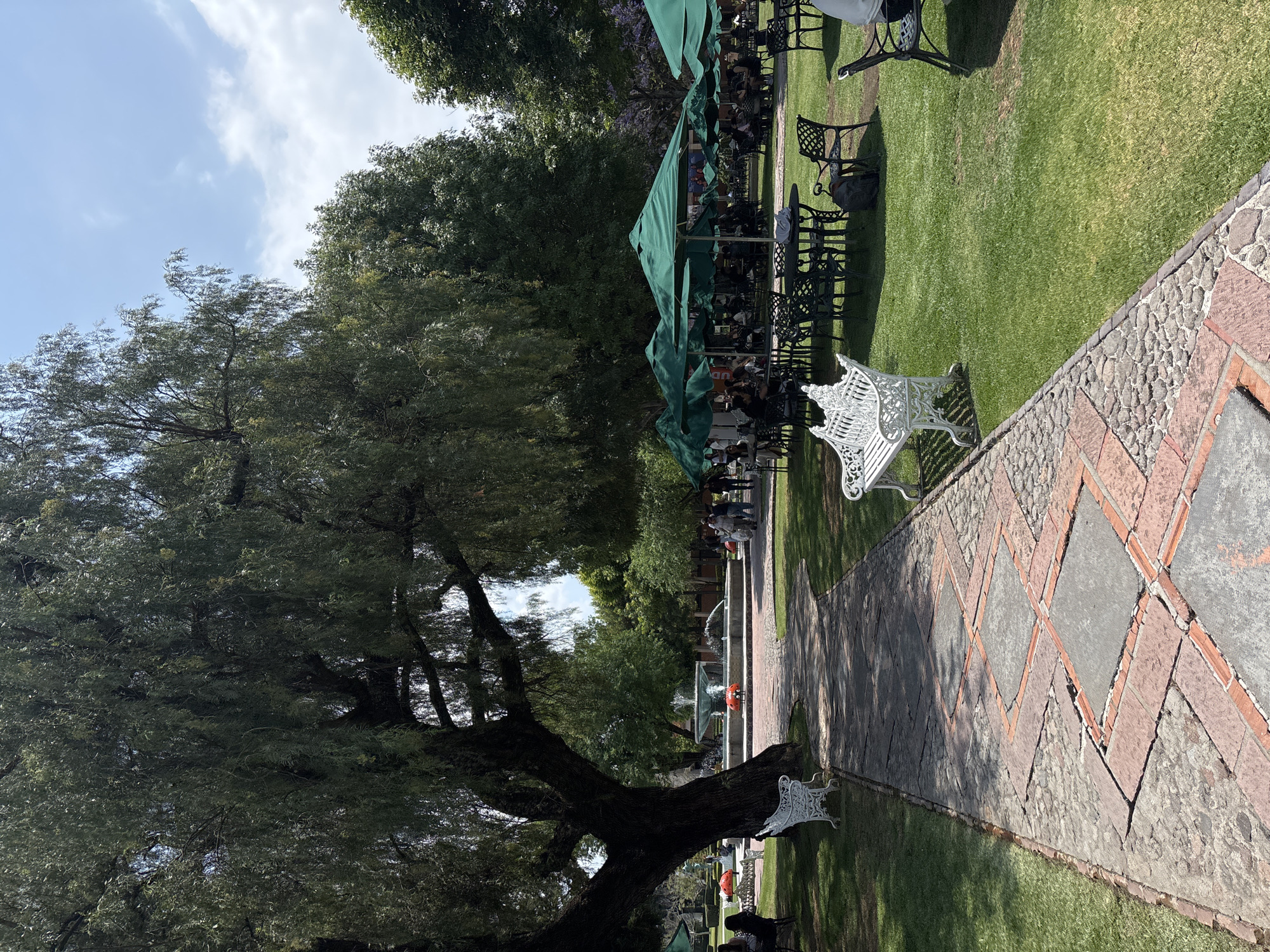 The image shows a park in San Andrés Cholula, Mexico. In the center is a brick and stone walkway leading towards a water fountain in the distance. A large tree dominates the left side of the frame. To the right, a lawn with green umbrellas and tables suggests a restaurant or outdoor seating area. White benches are placed along the walkway. The sky is visible through the trees, with a few scattered clouds. People are seen dining under the umbrellas.