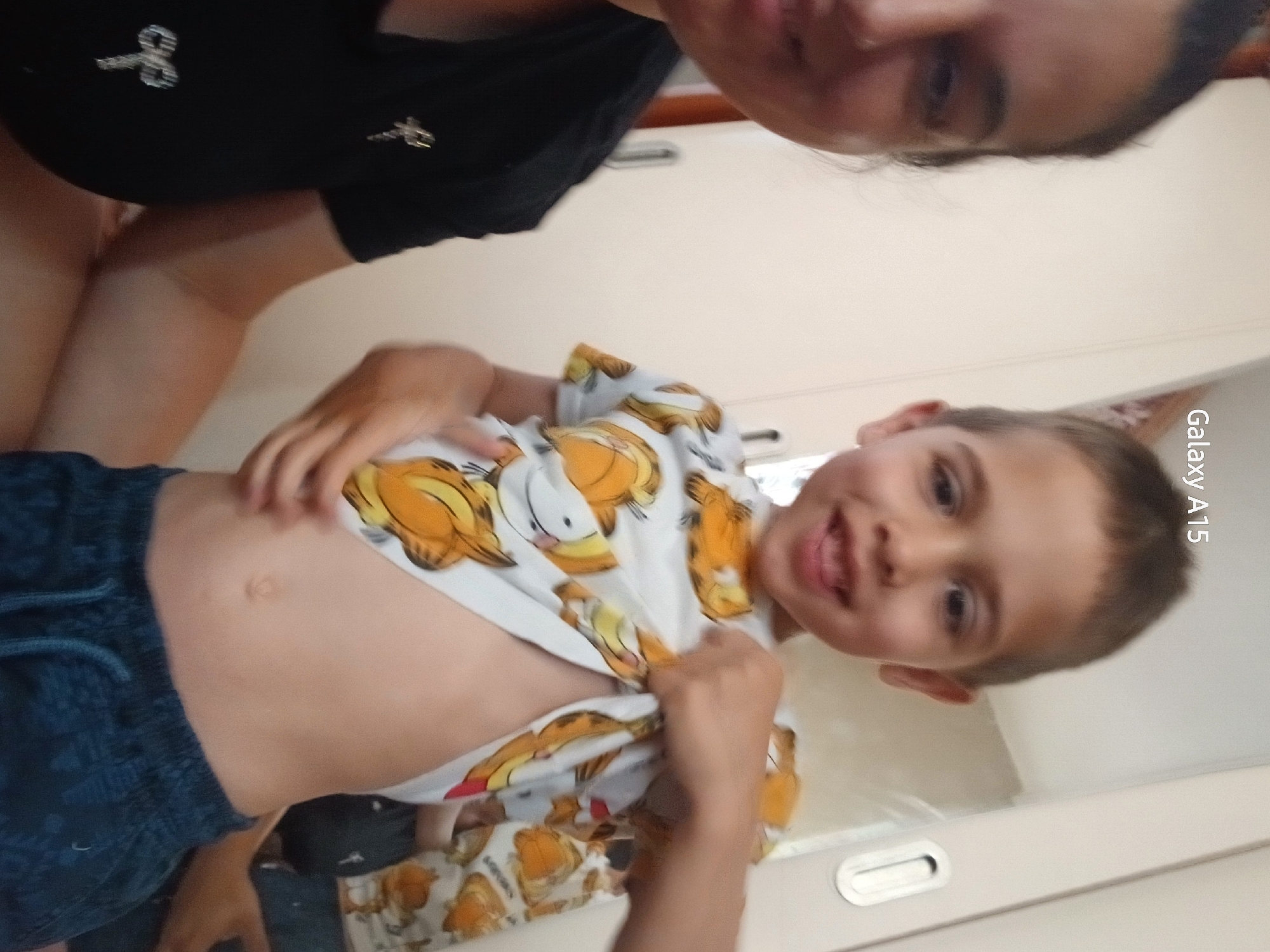 An indoor, vertically oriented close-up photograph captures a playful interaction between an adult and a young child, likely a boy, in Obrochishte, Bulgaria. The image is taken from a low angle, looking upwards at the subjects, which gives them a tilted orientation within the frame.
At the top of the photograph, an adult is partially visible, wearing a black top with small, shiny silver or white decorative elements, possibly bows or star shapes. The adult's face is partially seen on the upper right, looking towards the camera with a gentle expression.
The main subject is a young boy, positioned prominently in the center. He has short, light brown hair and is smiling broadly at the camera. He is wearing a white t-shirt adorned with multiple orange cartoon cat characters, resembling Garfield, and dark blue shorts. The boy is actively lifting his t-shirt with both hands, playfully revealing his bare stomach and belly button.
The background consists of light-colored walls or furniture, possibly cabinets or a door. A silver or white recessed handle is visible on the right side of this background element. The lighting suggests an indoor setting during the day or in a well-lit interior. The visible text "Galaxy A15" is present vertically along the right edge of the image, indicating the camera device used to capture the photo. The overall scene conveys a cheerful and informal atmosphere.