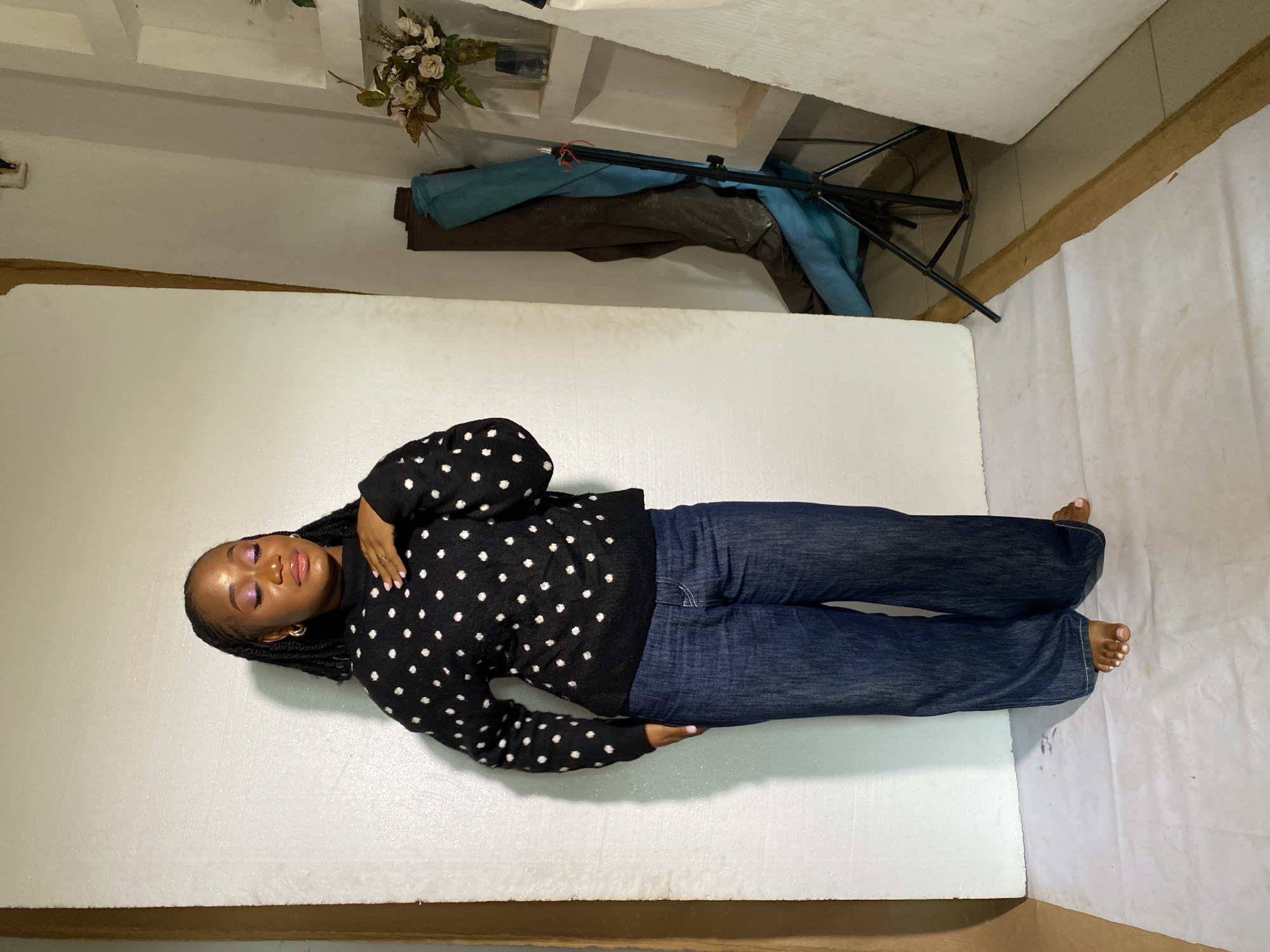 A young Black woman stands barefoot in a studio, wearing a black, high-necked sweater with white polka dots and wide-leg dark wash denim jeans. She has her eyes closed and her left hand resting on her chest. Her hair is styled in braids that fall to her shoulders. The background is a plain white wall. To the right, a tripod with a light stand is visible, along with rolled-up fabric and a vase of flowers on a shelf. The floor is covered with a white sheet. The image is set in a studio in Bukuru, Nigeria.