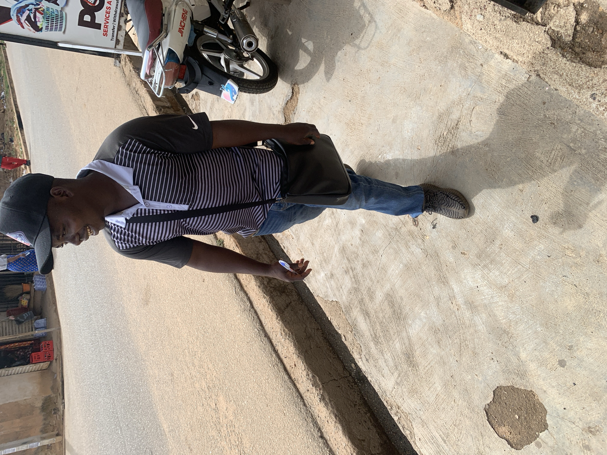 The image depicts a man standing on a concrete surface, likely a sidewalk, in an urban setting. He is wearing a black baseball cap, a striped short-sleeved shirt with a white collar, blue jeans, and patterned shoes. He has a black shoulder bag. In his right hand, he holds a small blue and white object, possibly a pen.
To the left, a street is visible with shops or stalls lining the side. There are various items for sale, including what appears to be clothes and other goods.
To the upper left, a sign is partially visible, indicating business services with the word "SERVICES" prominently displayed. A parked motorcycle is also visible.
Based on the visual context and reference to Jos, Nigeria, the environment is likely a commercial area in Jos. The scene suggests a casual interaction, with the man standing and holding the item, possibly engaged in a transaction or conversation.