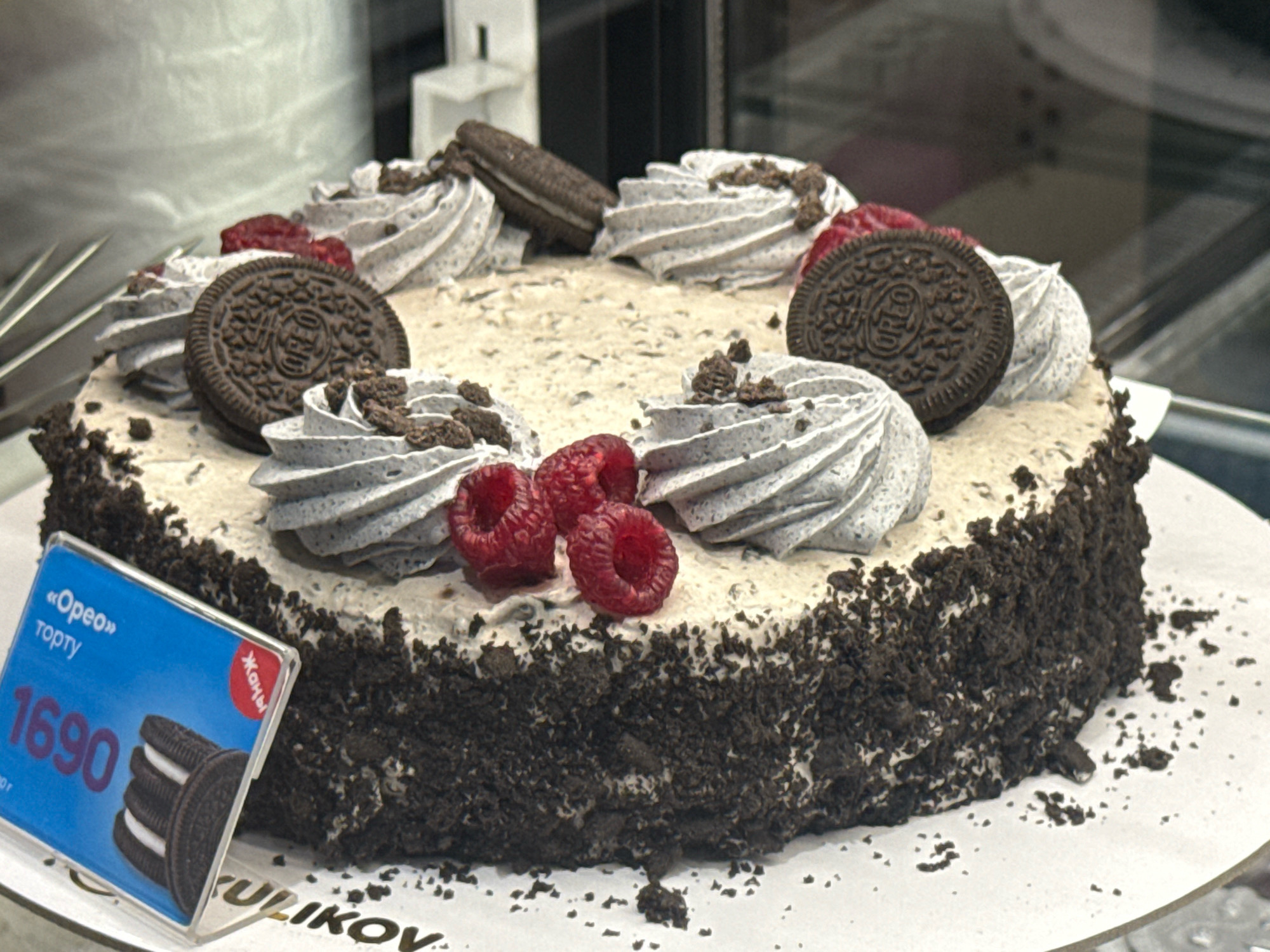 The image shows a close-up view of an Oreo cake. The cake is round and has a thick crust of crushed Oreo cookies. The top of the cake is covered in a light-colored frosting with dollops of grey frosting, decorated with Oreo cookies and fresh raspberries. A price tag for the cake, with "Oreo" written on it in Russian and the price "1690," is visible in the lower left corner. The cake is placed on a white cake stand. In the background, out of focus, there are shelves and some bakery display cases, suggesting the image was taken in a bakery or confectionery shop. The lighting is indoor and appears to be from artificial sources. The overall scene is a still life of a dessert item. No individuals are visible in the image.
