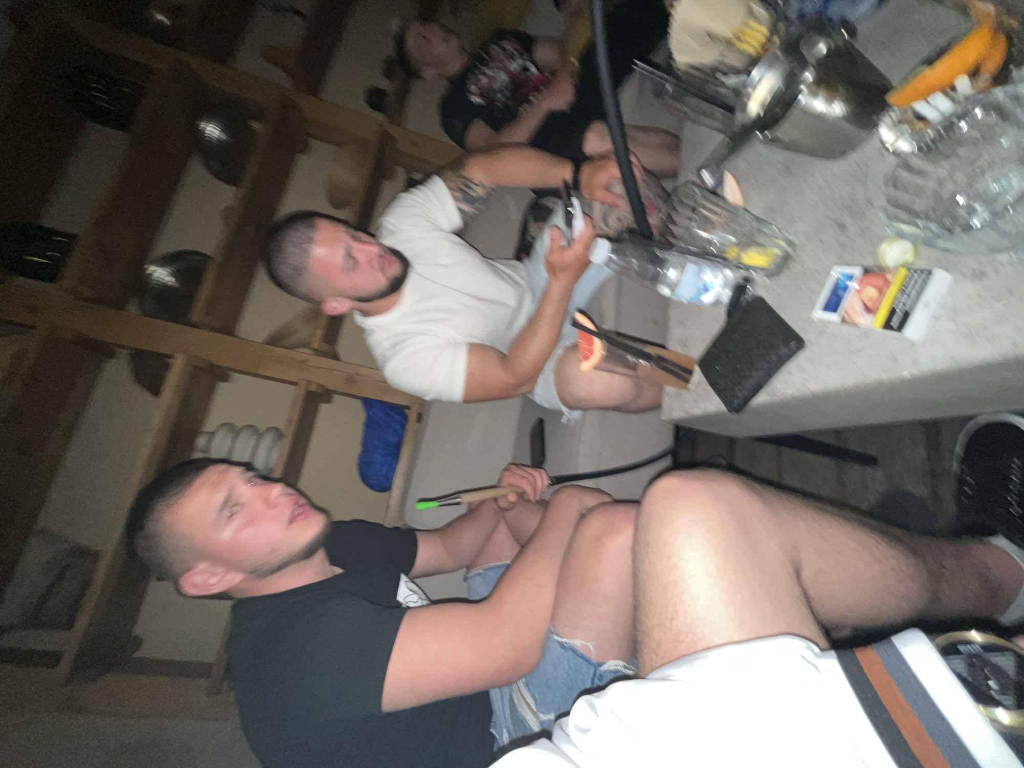 The image depicts an indoor scene where several individuals are gathered. Two men are prominent in the foreground; one wearing a black T-shirt and denim shorts, and the other wearing a white T-shirt. An individual in the background wears a black T-shirt with red/white graphics. A table nearby has several objects scattered, including a pack of cigarettes, a metallic container, and a glass carafe. The location, Burgas, Bulgaria, cannot be identified from the image.
