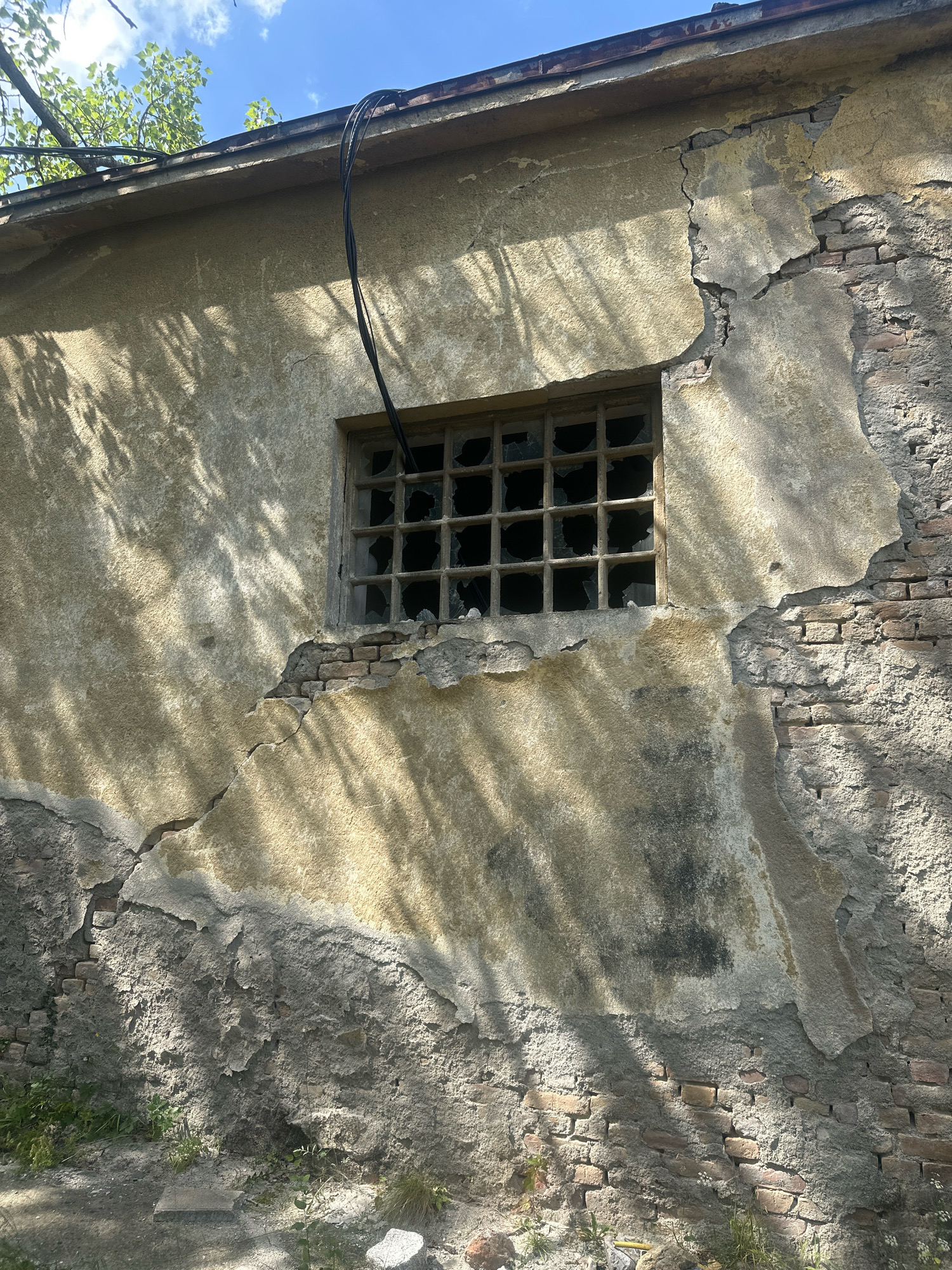 The image shows the exterior of a damaged building with peeling plaster, exposed brickwork, and a broken window. The building appears to be old and in disrepair, with cracks and crumbling sections on the wall. The window is barred and the glass panes are shattered. Black electrical cables are visible running along the top of the building and down towards the window. Tree branches with green leaves are seen against a blue sky with some clouds. The ground in front of the building is covered with dirt, grass, and some debris. The scene is likely located in an outdoor, possibly urban or suburban environment, given the presence of a building and electrical infrastructure. The overall impression is one of decay and neglect.