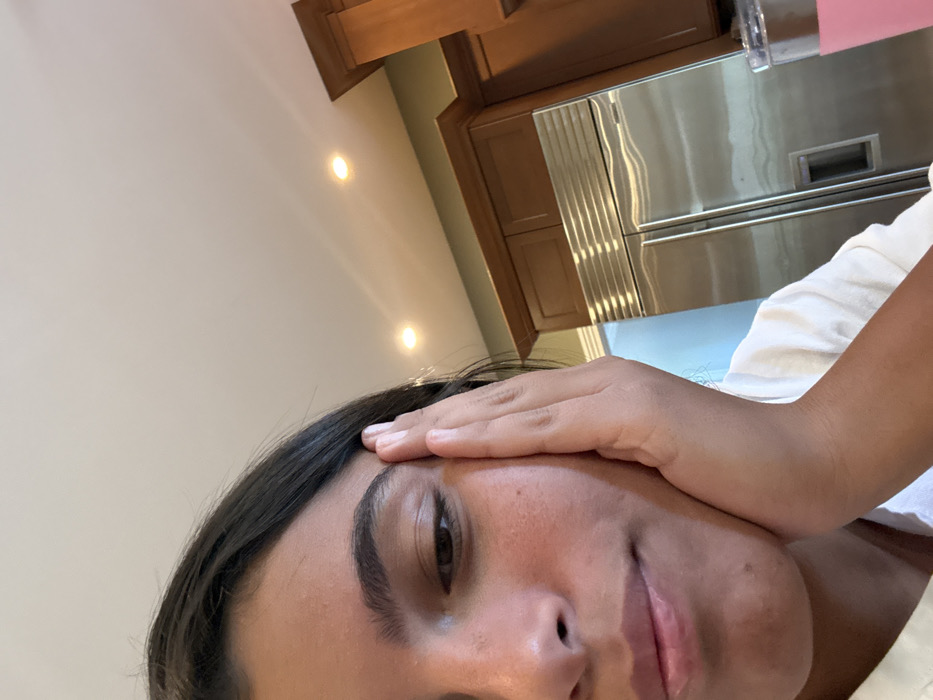 The image depicts an indoor kitchen setting. A single individual, appearing to be a young female with dark hair and medium skin tone, occupies the lower-left portion of the frame. Her left hand is resting against the left side of her face, near her temple and cheek. She is wearing a light-colored top, and her visible eye is cast downwards, indicating a neutral or contemplative expression.
In the background, to the right of the individual, a large stainless steel side-by-side refrigerator is visible, featuring vertical handles and horizontal ventilation grilles. Above and to the right of the refrigerator, light brown wooden kitchen cabinets are mounted on a wall painted light green. The ceiling is white, with two illuminated recessed spotlights visible. In the bottom-right corner, a clear tumbler containing a pink liquid is partially visible. A bright, undefined light source or reflection appears to the left of the refrigerator. The specific location within Princeton, United States, cannot be determined from the visual information presented.