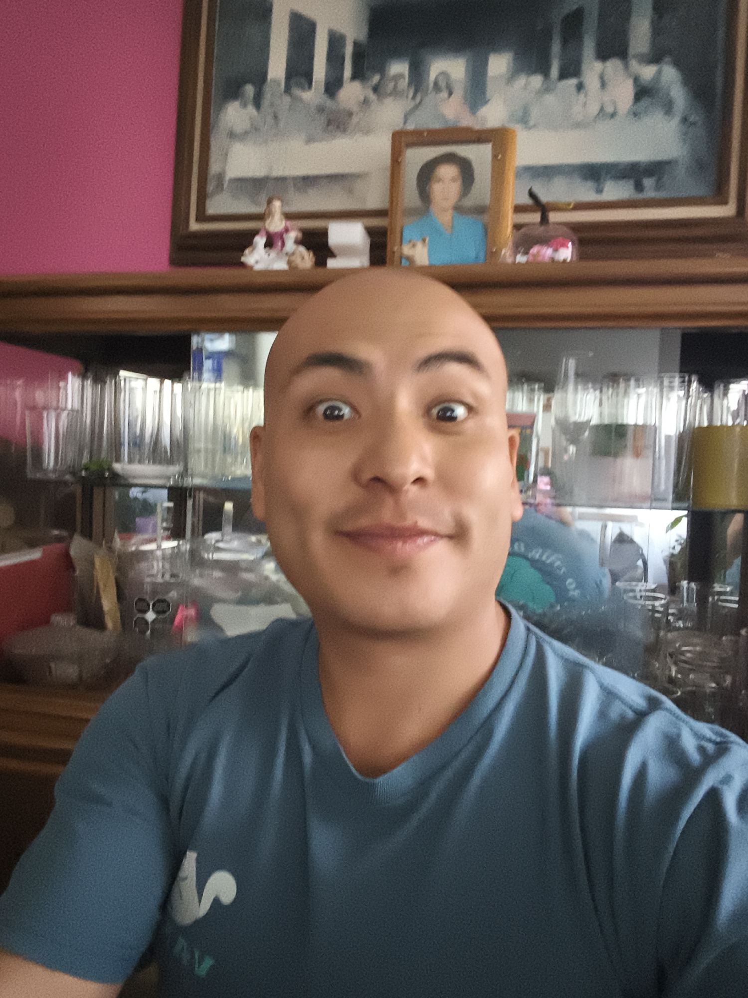A close-up selfie of a bald man with wide eyes and a slight smile, wearing a blue t-shirt. Behind him is a wooden cabinet with a mirrored back, reflecting numerous clear glasses. On top of the cabinet are two framed pictures and a small figurine of a woman. The wall behind the cabinet is pink. The location is likely a home interior in Mexico City, Mexico.