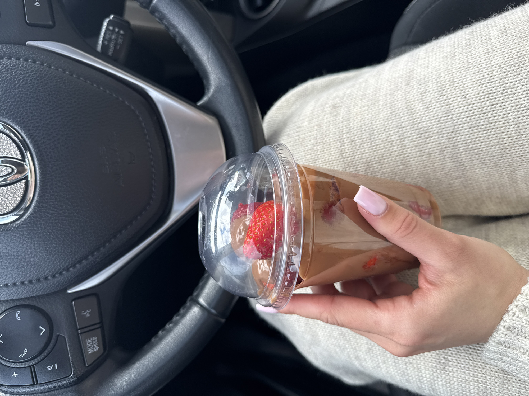 A close-up shot inside a car in Kyiv, Ukraine, shows a person's right hand holding a clear plastic cup. The cup contains a brown, creamy substance, likely chocolate or Nutella, with several pieces of cut fresh strawberries visible through the plastic and beneath the domed lid. The person has manicured nails with a pink and white French tip design and is wearing a light-colored, possibly beige, knitted garment on their lap or leg.
The background reveals the interior of a car, specifically the steering wheel and part of the dashboard. The steering wheel is black, appearing to be leather, with silver accents. The Toyota logo is prominently displayed in the center of the steering wheel, which also bears the text "SRS AIRBAG". To the left of the center, various control buttons are visible on the steering wheel, including those for phone calls and navigation. One button clearly reads "MODE HOLD". Behind the steering wheel, a stalk with controls labeled "RES +" and "SET -" is partially visible. The lighting suggests it is daytime.
