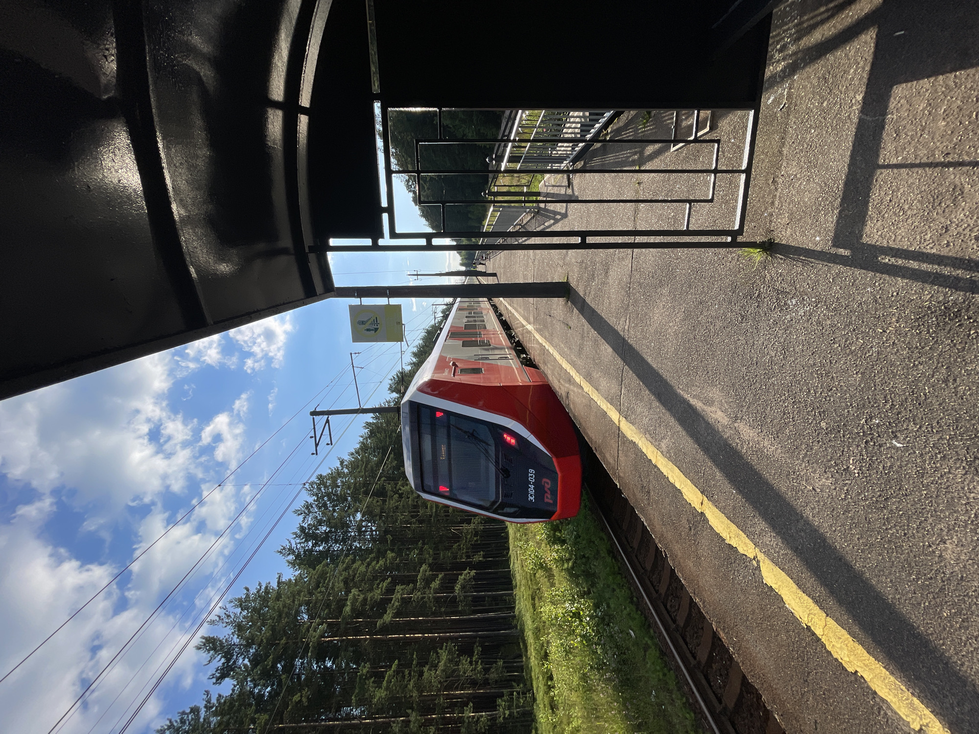 A red and white passenger train is stopped at a rural train station platform. The train is positioned on the left side of the image, with its front facing the viewer. The platform, made of asphalt, runs along the right side of the train. A bright yellow line is painted along the edge of the platform. In the background, a dense forest of pine trees lines the opposite side of the train tracks. Above, a bright blue sky is dotted with white clouds. The right side of the image is dominated by a dark, shadowed structure, possibly a shelter or overhang, with a black metal gate visible in the foreground. The scene appears to be taken in the city of Lebyazhye, Russia, given the context provided.