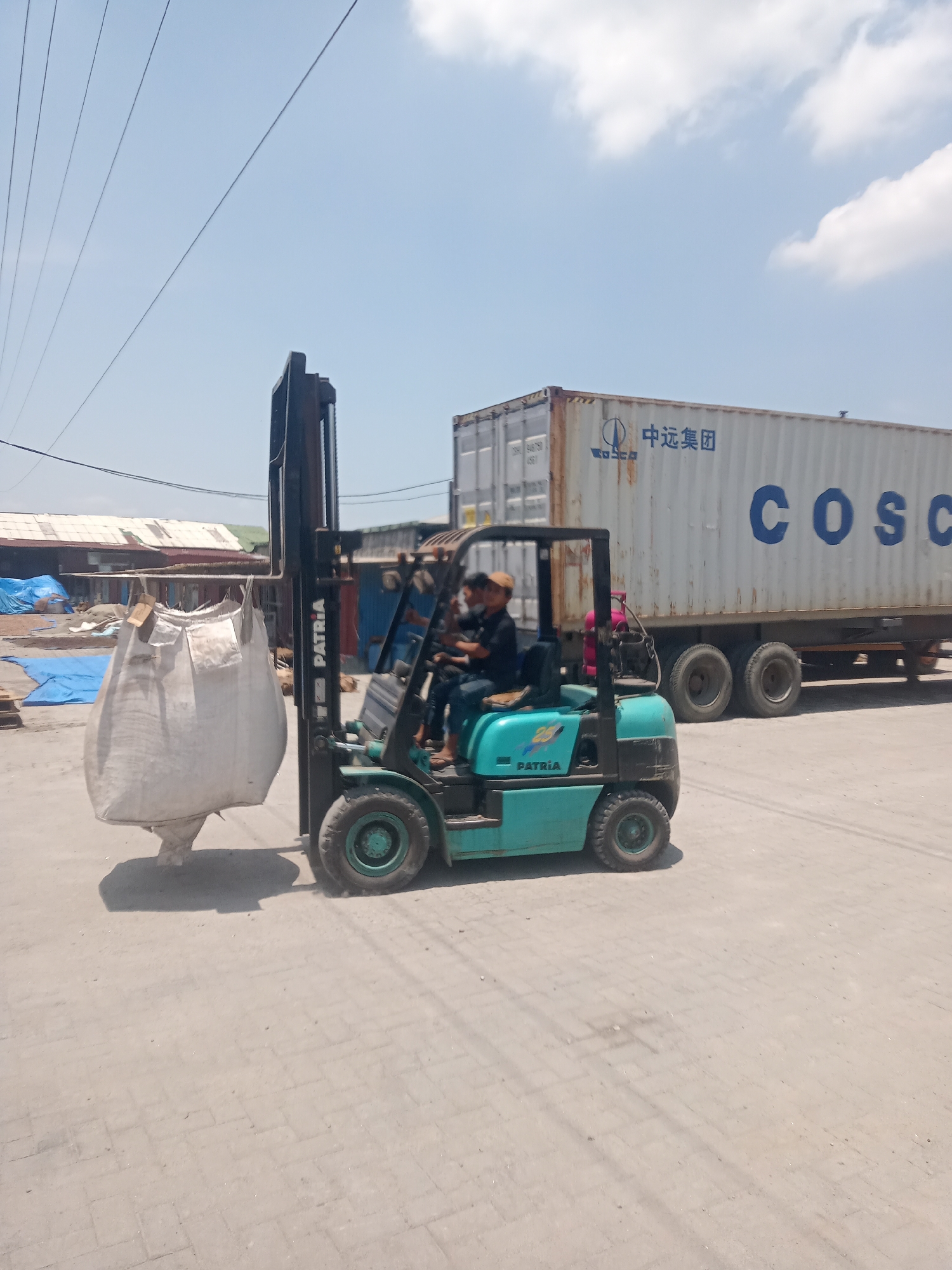 A teal and black forklift with "PATRIA" branding operates on a paved outdoor area, likely a loading dock or industrial yard in Sunggal, Indonesia. The forklift is lifting a large, white, woven bag, suspended by its handles. Two men are seated on the forklift; one is operating it, and the other is a passenger. Behind the forklift is a white shipping container with blue lettering that reads "COSCO" and Chinese characters. The container is attached to a truck with multiple axles. In the background, several buildings with corrugated metal roofs are visible, along with blue tarps covering materials on the ground. Overhead, power lines stretch across a bright blue sky with scattered white clouds. The ground is a mixture of paved surfaces and dirt.