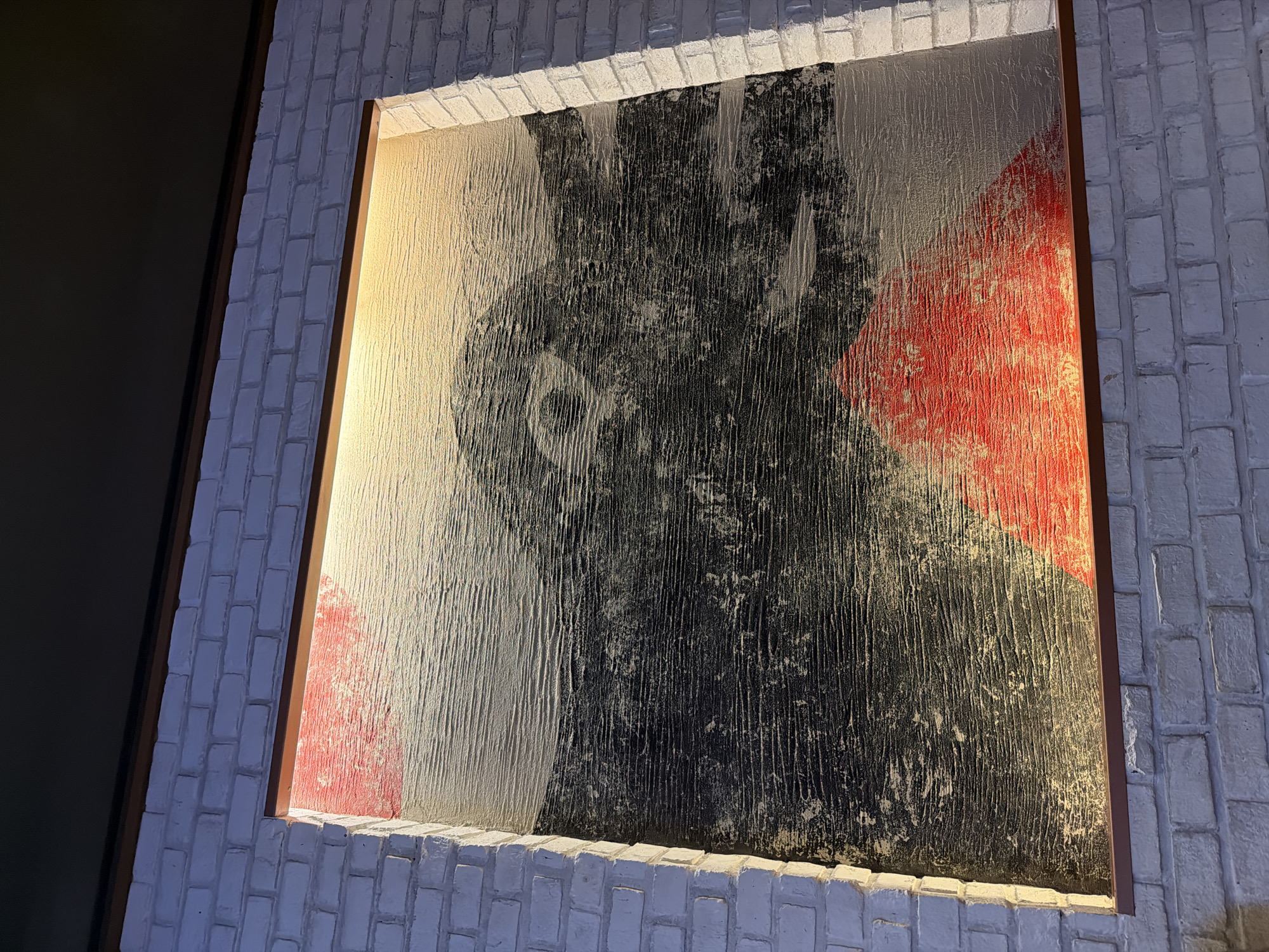 The image shows a framed abstract painting on a white brick wall in a location identifiable by the city of บ้านบางรักษ์, Thailand. The painting features vertical lines of white, black, and red paint. The frame is a thin, brown color. The wall is constructed of painted white bricks. The lighting highlights the texture of both the painting and the wall. A dark area is visible on the left side of the image, suggesting an adjacent wall or object.