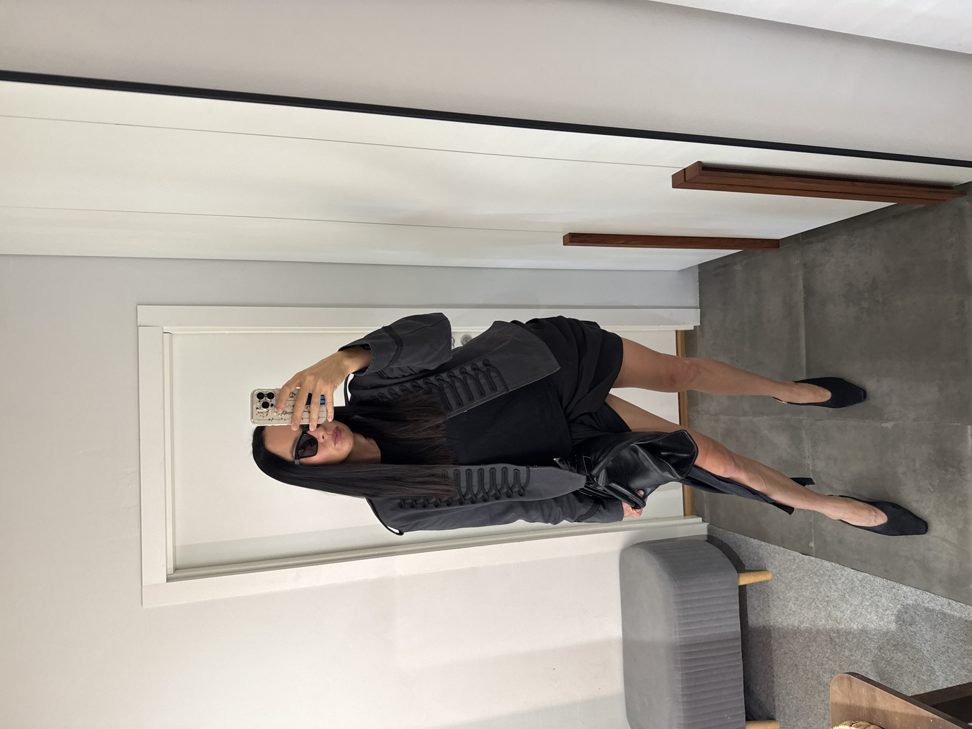 A woman is taking a mirror selfie indoors. She is wearing a black t-shirt, a dark gray jacket with decorative braiding, black shorts, and black pointed-toe heels. She has on dark sunglasses and is holding a black leather clutch bag. The background consists of a white door, a gray upholstered bench, a wooden table, and a white closet or wall. The flooring appears to be concrete or a similar material.