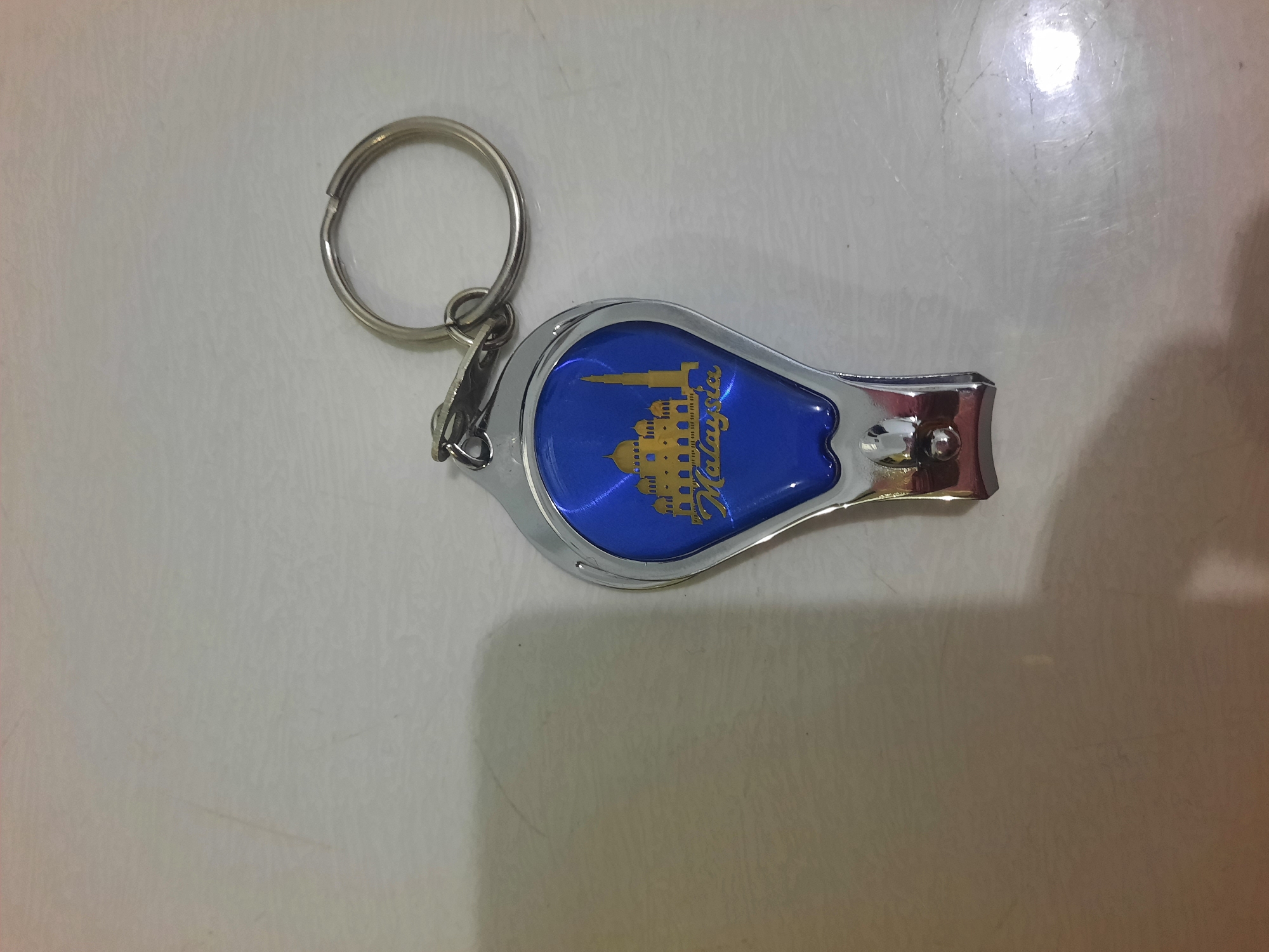 A keychain featuring a nail clipper with a blue, teardrop-shaped insert is pictured on a light-colored, textured surface. The insert has a gold silhouette of a mosque with domes and a minaret, and the word "Malaysia" written in gold script below the silhouette. A silver keyring is attached to the top of the nail clipper. The surface appears to be a countertop or table, with faint lines and reflections visible. There is no indication of the city of Lhokseumawe, Indonesia in the image.