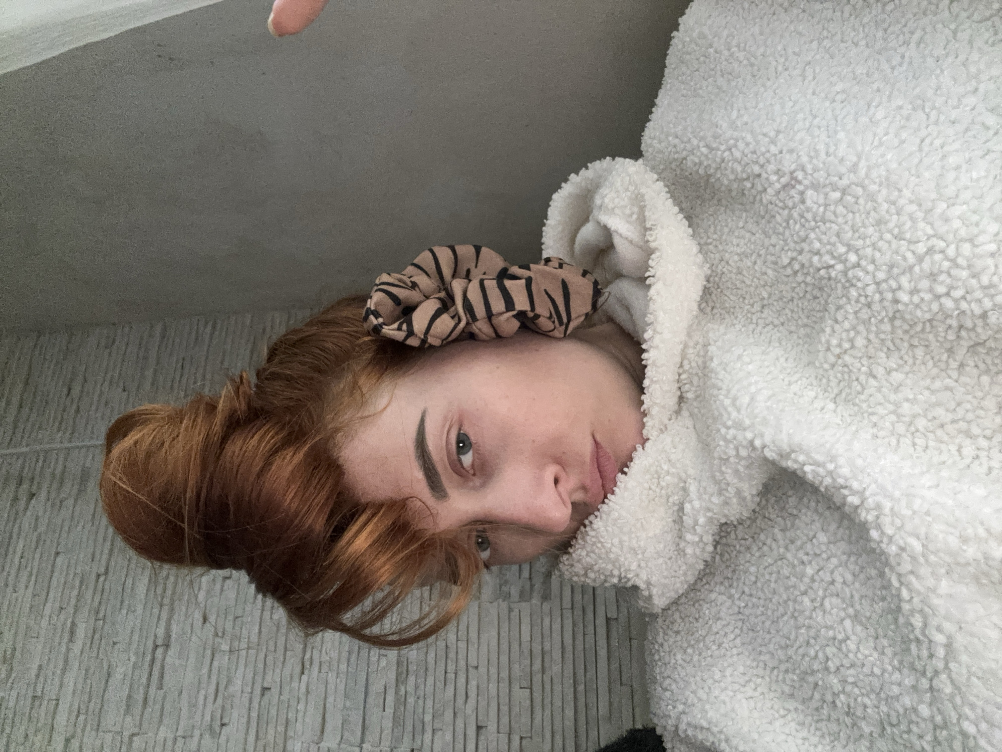 A woman with red hair styled in a bun and secured with a patterned scrunchie is photographed from an unconventional angle, tilting her head to face the camera. She wears a fluffy, white hooded garment, possibly a jacket or robe, with the hood pulled up around her face. The background features a wall with a vertical, stacked-stone pattern. The location is potentially Budva, Montenegro, but there are no specific details to confirm the location in the image.