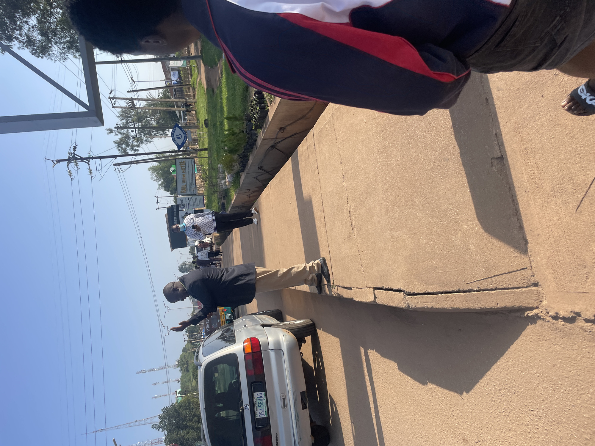 The image shows a street scene in Jos, Nigeria, with a man in a suit and khaki pants standing on a sidewalk, gesturing with his right hand raised. To his left, a silver minivan is parked. Behind him, a woman in a patterned top and pants is walking away from the camera. In the background, there are buildings, trees, power lines, and communication towers. The sky is clear and blue. A "Rail View Hotel" sign is visible on the left side of the image.