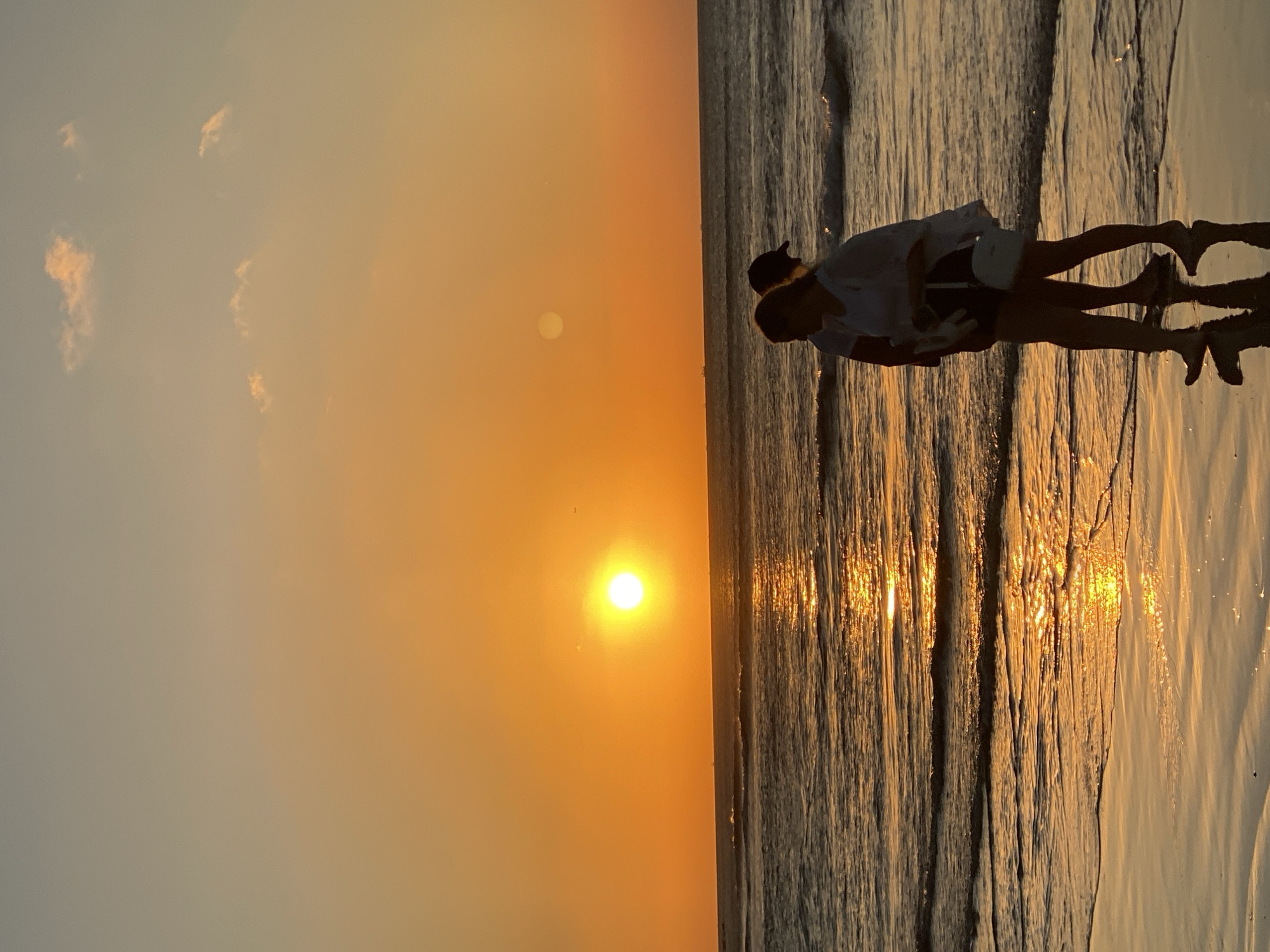 The image captures a stunning sunset at a beach in Kuta Selatan, Indonesia. The sun, a brilliant orange sphere, is positioned low on the horizon, bathing the scene in warm, golden light. The sky transitions from a fiery orange near the sun to a softer, muted grey-blue further away, with a few faint clouds scattered across the upper portion.
Two figures are silhouetted against the radiant backdrop of the setting sun. They appear to be standing at the water's edge, with their backs partially turned, facing the horizon. One figure is taller, possibly an adult, while the other is shorter, suggesting a child or a second adult, standing closely together. Their forms are dark and distinct against the bright sky and the shimmering reflections on the water.
The ocean surface is gently rippled, reflecting the intense golden light of the sun in long, broken streaks that extend from the horizon towards the foreground. The overall scene conveys a sense of peace and natural beauty, characteristic of an evening on a tropical beach. There is no visible text in the image.