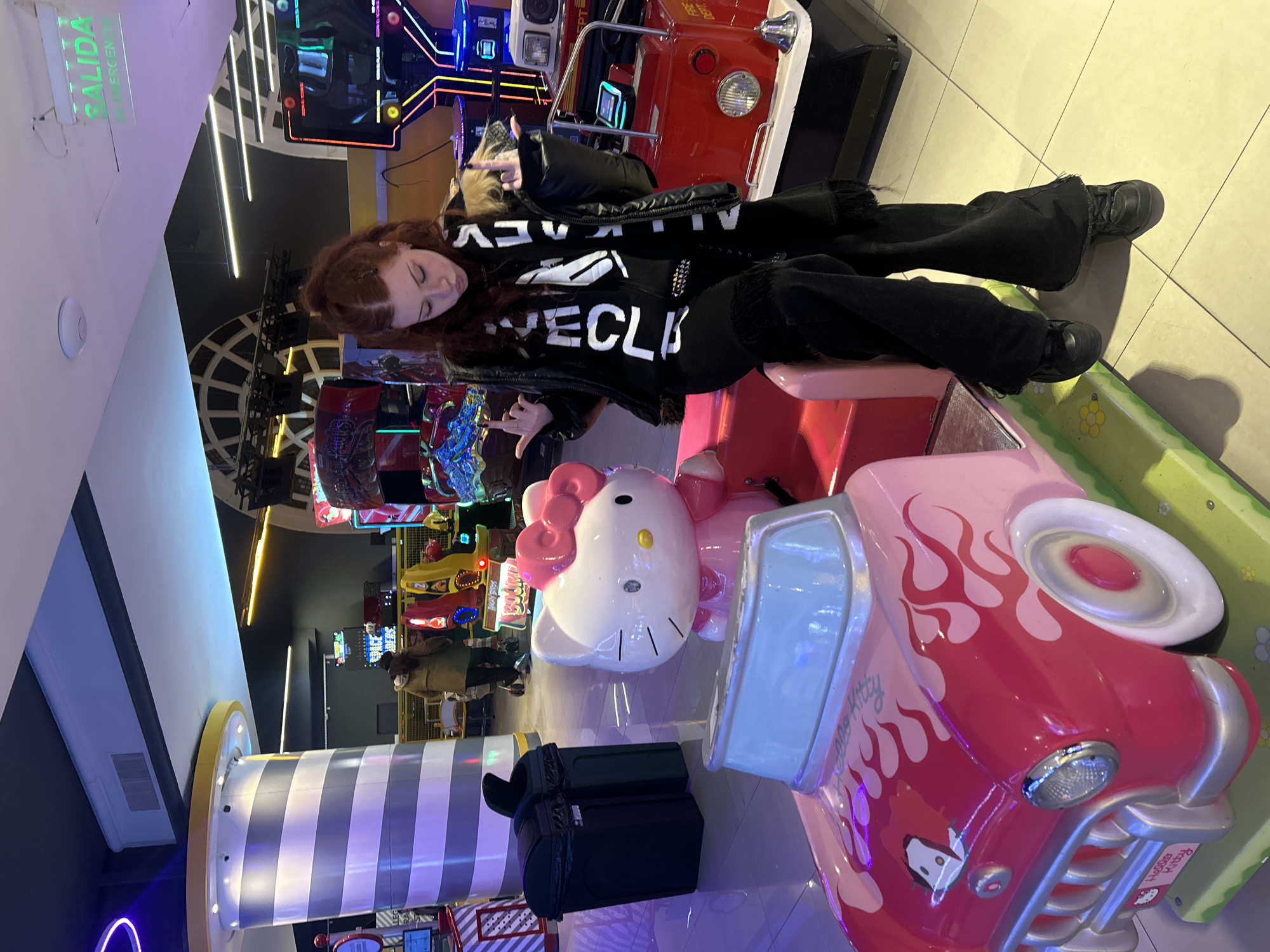 A young female with red hair sits on a pink Hello Kitty themed amusement ride at an indoor arcade. She is wearing black pants, a black jacket, and a black and white scarf. The arcade appears to be in Buenos Aires, Argentina, given the "SALIDA" (Exit) sign in Spanish. The background shows various arcade game machines and a large striped pillar.