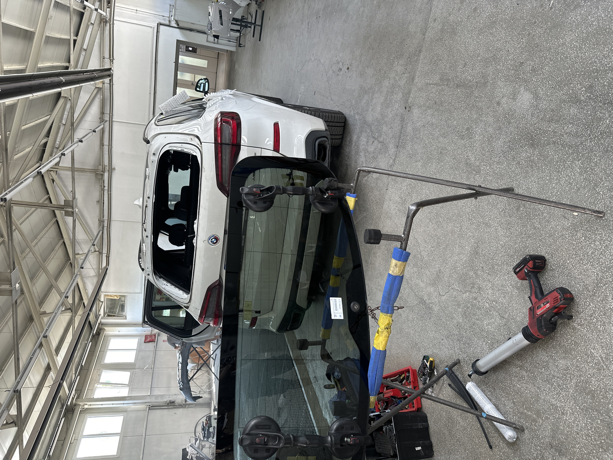 The media file captures an indoor automotive repair or glass replacement workshop. The primary subjects are a white SUV, a large piece of automotive glass, and various tools.
The white SUV, identified as a BMW by the visible logo on its tailgate, is undergoing repair. Its rear bumper has been removed, and other parts may also be detached, indicating ongoing work. A small white cleaning pad rests on its roof.
In the foreground, a large piece of automotive glass, likely a rear window or windshield, is propped up and ready for installation. It has two large black suction cups attached for handling. A white sticker with a barcode and the text "BMW CAR" is visible on the glass.
Surrounding the car and glass are several tools and equipment. A red cordless caulk gun, along with several white tubes of sealant or adhesive, lies on the concrete floor. A red toolbox, partially visible, contains various hand tools such as wrenches and screwdrivers. Several metal support stands, some padded with blue and yellow material, are positioned around the work area, likely to assist in holding the glass or car parts.
The setting is a well-lit garage or workshop with a concrete floor. The walls are light-colored, and the ceiling features an industrial structure of metal beams and supports. Multiple windows on one wall allow natural light to illuminate the space, suggesting it is daytime. A person is partially visible in the background, possibly through the car's open door or as a reflection, indicating active work.
The image depicts a specific stage of car repair, focusing on the replacement of automotive glass, with all the necessary tools and the vehicle in a state of disassembly.