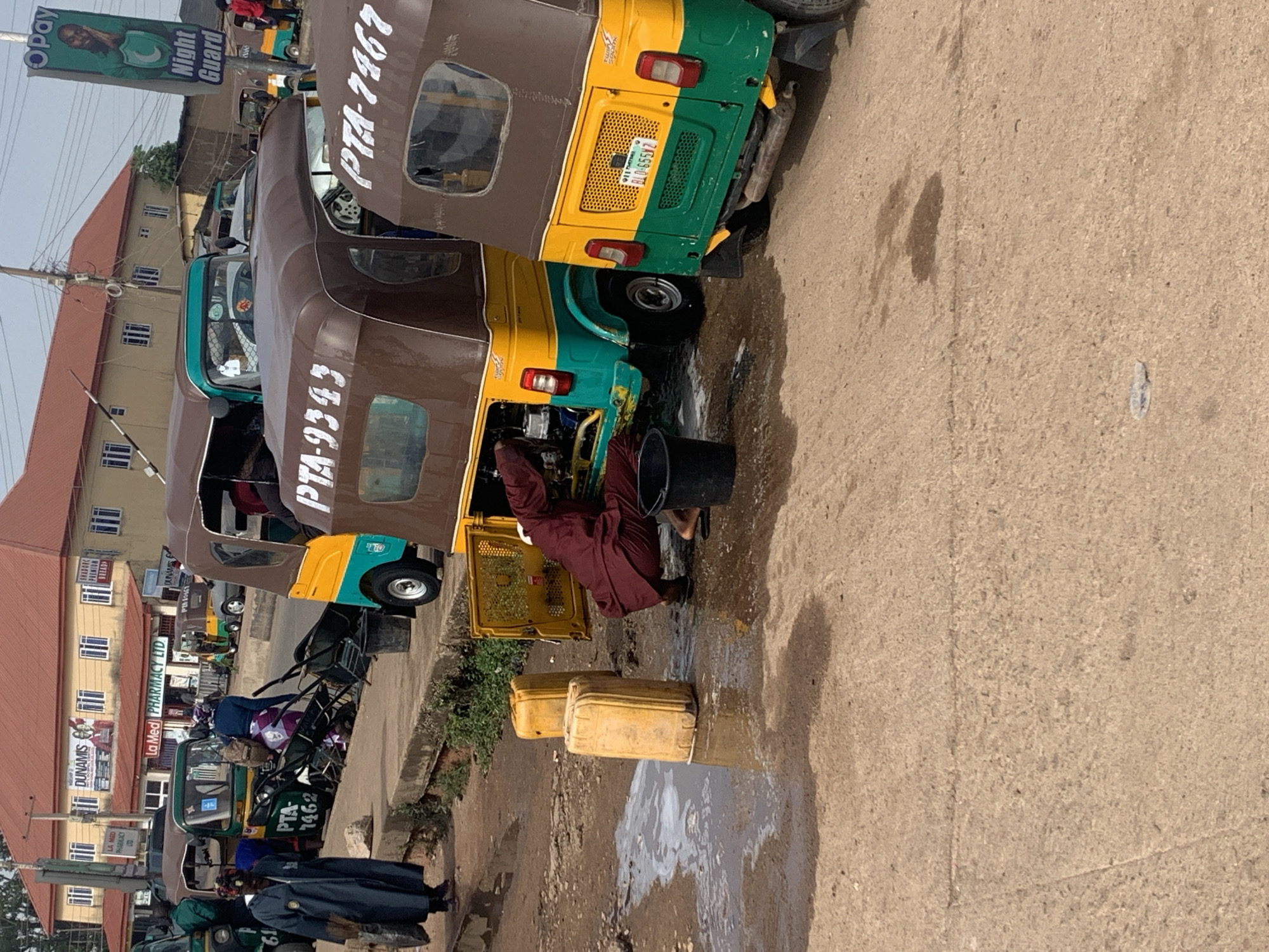 The image depicts a scene in Gura Top, Nigeria, where several auto-rickshaws, also known as Keke Napep, are parked. In the foreground, two yellow plastic jerry cans are positioned on the wet ground, suggesting recent activity. A person, dressed in a maroon tunic and trousers, is crouched beside an open-hooded auto-rickshaw, appearing to be working on its engine. The auto-rickshaw is partially disassembled, with its engine exposed. A black bucket is located near the person. To the right, another auto-rickshaw with the license plate "BLO-655VZ" is visible, its rear end facing the viewer. Further back, additional auto-rickshaws are parked, and a woman in a blue robe is standing on the sidewalk. A building with a red roof and the signage "La Med Pharmacy LTD" is in the background, along with advertisements and other businesses. The environment appears to be an urban setting with a paved road and sidewalk.