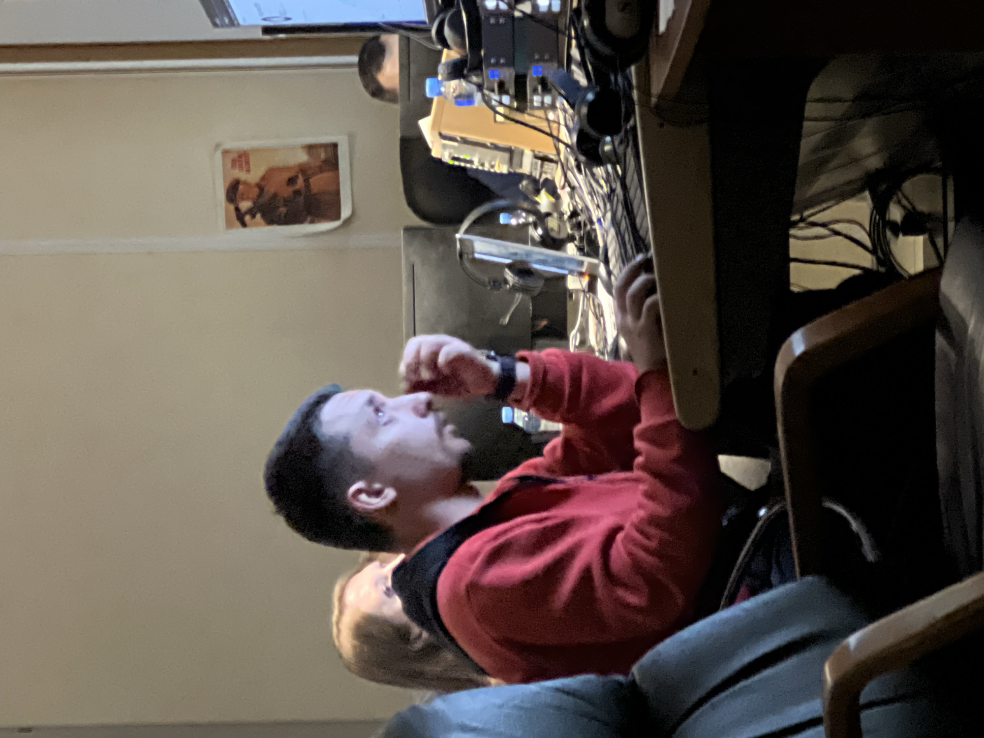 A man wearing a red sweater is seated at a desk with electronic equipment. Another person is seated behind him. A poster is visible on the wall. The location is not discernible from the image, therefore Khimki, Russia, cannot be confirmed.