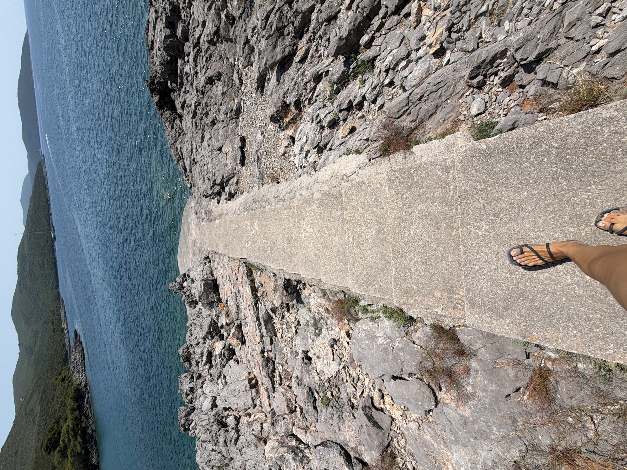 The image shows a paved walkway leading down towards a body of blue water. The walkway is flanked by rough, rocky terrain on both sides. In the foreground, a person's legs and feet are visible, wearing black sandals and walking down the path. The water appears to be calm, with a few small waves. In the distance, there are hills covered in green vegetation, and a sailboat can be seen on the water. The sky is clear and blue. The scene depicts a scenic coastal environment, likely a pathway to access the water in Bigova, Montenegro.