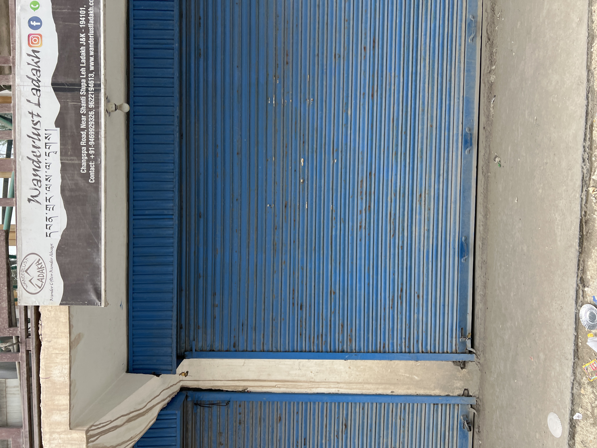 The image shows a storefront in Leh, India, with its blue metal shutters down. The shutters are worn and show signs of rust in places. A sign above the shutters reads "Wanderlust Ladakh" with social media icons and Tibetan script. Below the sign, contact information is displayed: "Changspa Road, Near Shanti Stupa Leh Ladakh J&K - 194101, Contact: +91-9469929326, 9622194613, www.wanderlustladakh.com". A single lightbulb hangs from the underside of the awning above the shutters. The ground in front of the shop is concrete and dirty, with some scattered trash including paper plates.