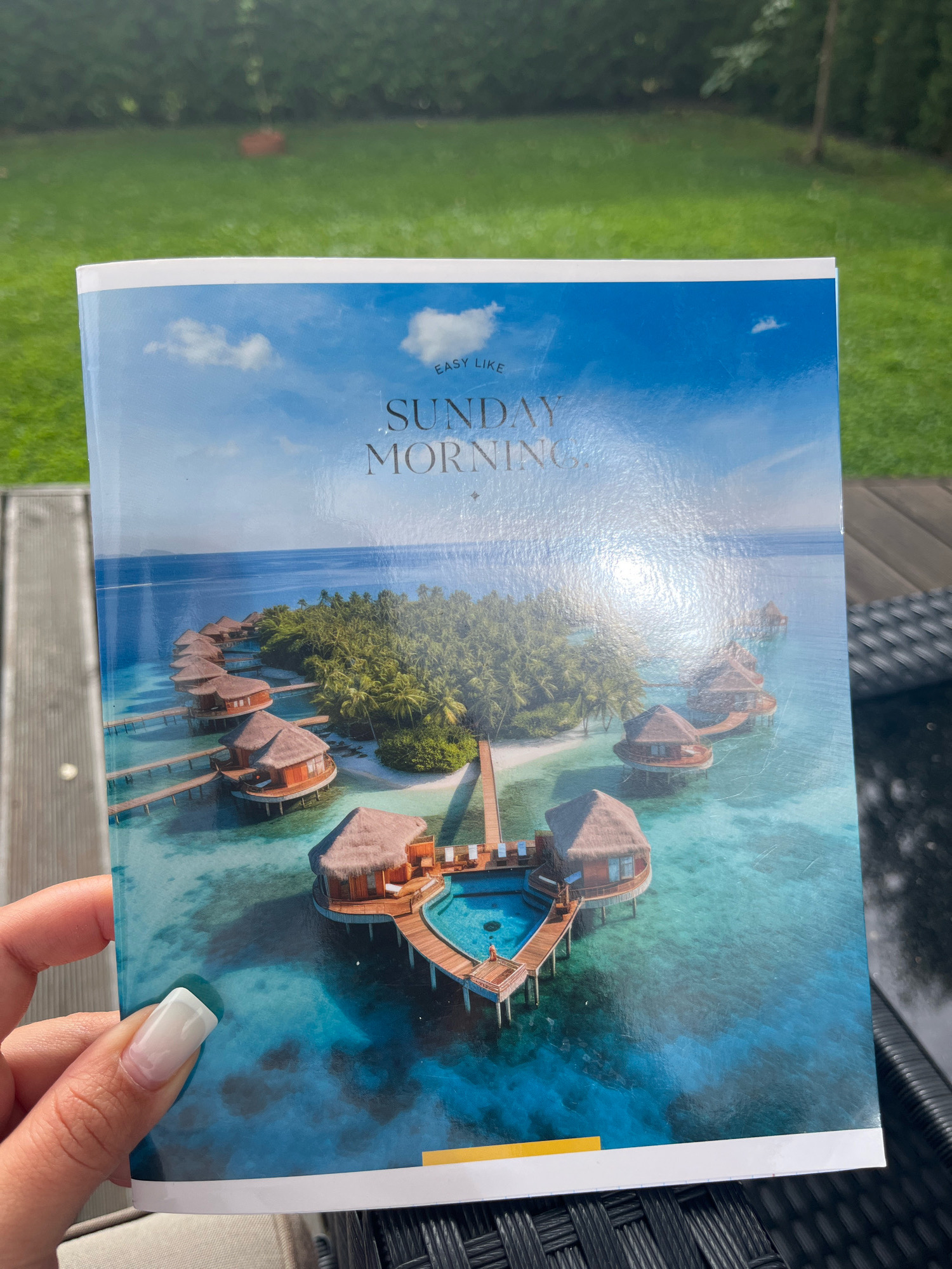 A hand with a white and pink ombre manicure is holding a brochure with a picture of overwater bungalows in a tropical location. The brochure features the text "EASY LIKE SUNDAY MORNING". The background shows a green lawn and what appears to be a wooden deck. The scene does not provide any information or context related to Haabneeme alevik, Estonia.