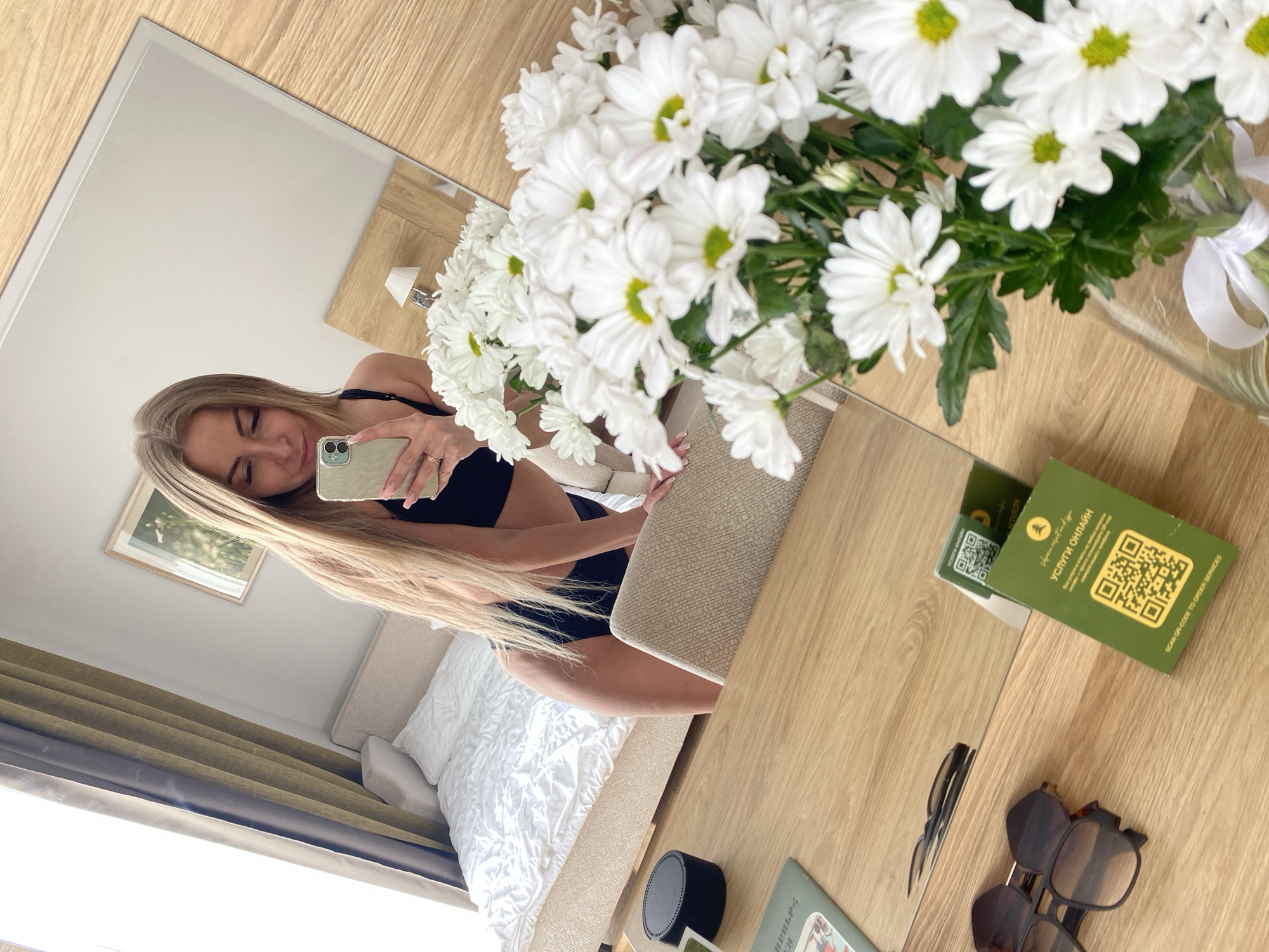A mirror reflects a blonde woman in a black bikini taking a selfie, with a bouquet of white daisies on a table. The table also holds a pair of sunglasses, a small black speaker, and a green card with a QR code labeled "Услуги онлайн" (Online Services). The reflection includes a bed, a framed picture, and a curtain. The location is indicated by the "Услуги онлайн" card, which is associated with services in Russia. Given the origin of the person that provided the photo and crops, it may be possible to deduce that the location is within the city of Tyumen, Russia.