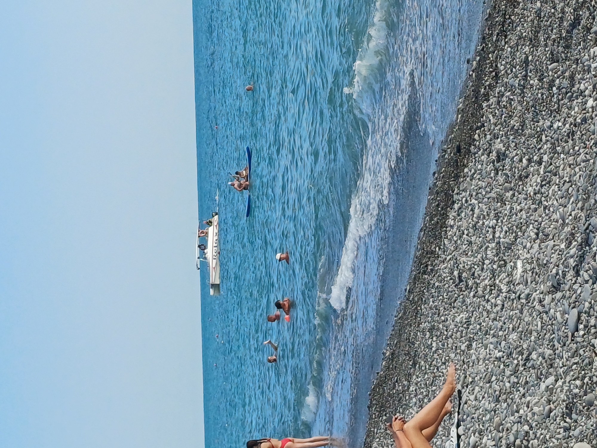 This is a vertical, eye-level, outdoor, medium shot of a beach scene in Sochi, Russia. The beach is covered in pebbles and the ocean is a vibrant blue with moderate waves. The sky is a clear, light blue, indicating it's likely daytime with good weather.
Several people are visible in the water. One person is on a paddleboard with another person, and a third person appears to be behind them. Further out in the water, another person is swimming. Nearer to the shore, a group of people are in the water, some appearing to be swimming and others standing. One individual in the water has their arm raised.
On the beach in the foreground, a person is lying on their back, wearing a red bikini. To the right of them, another person's legs are visible, also appearing to be sunbathing. A boat with the word "Relax" visible on its side is partially in the frame near the people in the water. There are also small, red buoys dotted in the water, possibly marking a swimming area.
The overall mood is one of leisure and recreation, with people enjoying a sunny day at the beach.