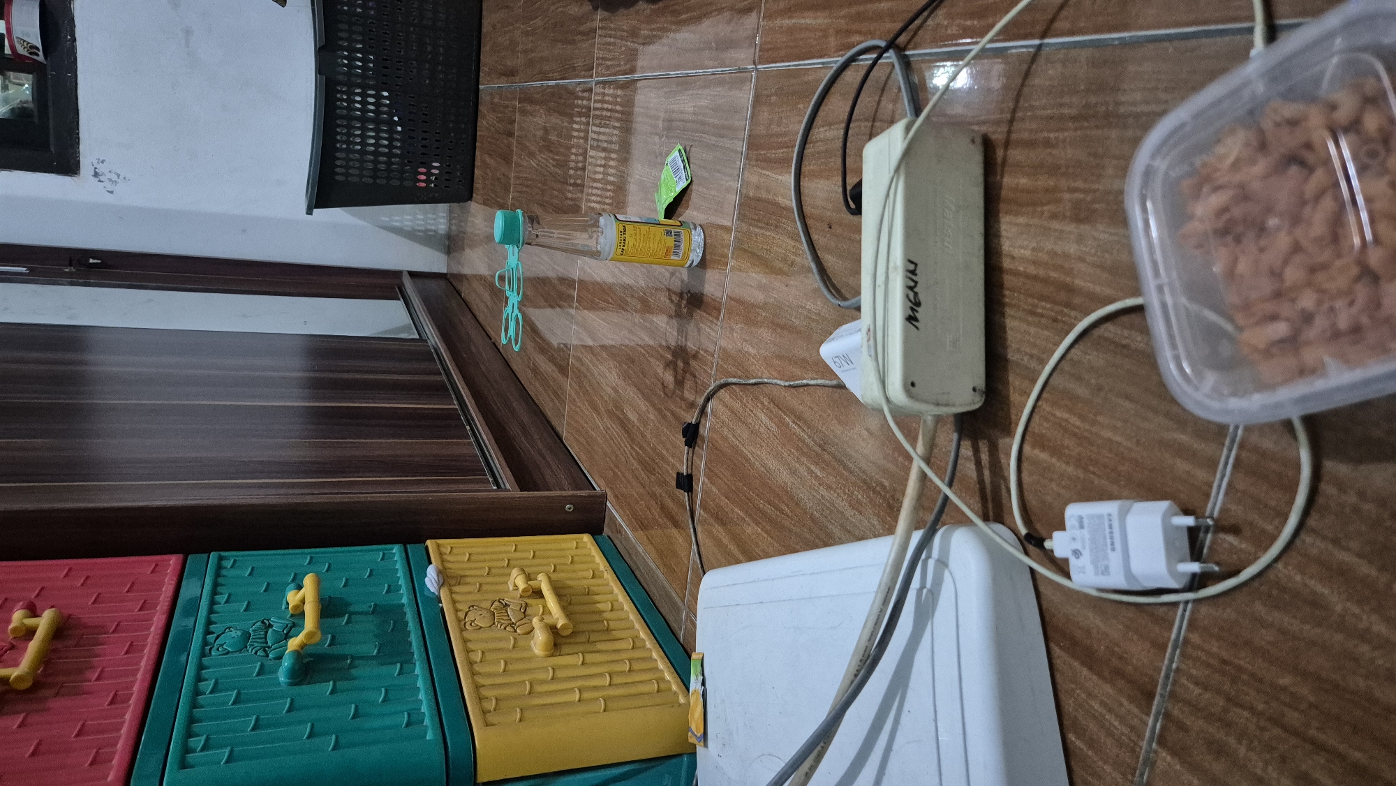 The image depicts a cluttered floor scene, likely in a residential setting in Karawang, Indonesia. In the foreground, a white appliance, possibly a scanner or printer, is connected by cables to a white power strip labeled "Menn". A white Samsung phone charger is plugged into the power strip, and its cable is coiled on the floor. To the right, a clear plastic container filled with what appears to be cooked pasta is visible. Further back, a stack of brightly colored plastic drawers, red, green, and yellow, stands against a wooden wardrobe. On the floor near the drawers, a small glass bottle of what seems to be oil with a green cap is present, along with a small green packet. In the background, a black plastic laundry basket is visible against a pale wall. The floor is tiled with a brown wood-grain pattern.