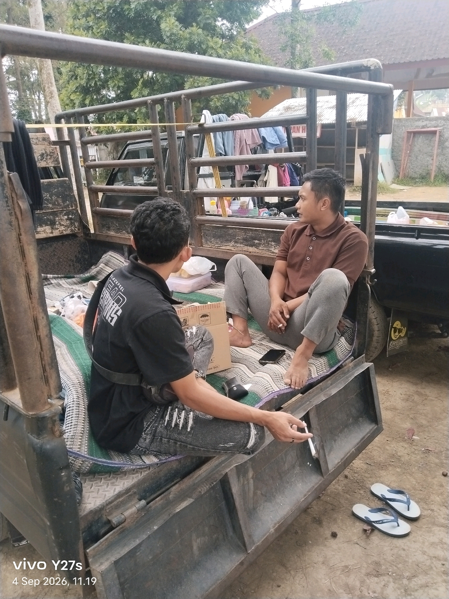 In the city of Maron, Indonesia, two men are sitting in the back of a pickup truck. The man on the left is wearing a black t-shirt and jeans, and the man on the right is wearing a brown polo shirt and grey pants. Both men appear to be in their late 20s or early 30s. The truck bed is covered with a woven mat, and there are various items in the truck, including clothes, boxes, and plastic containers. The men are sitting and talking, and the man on the right is holding a cigarette. The background shows trees and a building. The overall scene appears to be casual and relaxed.