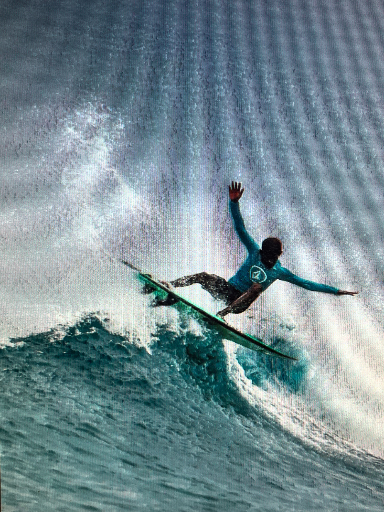 In an outdoor, natural environment, a surfer is in mid-air, having launched off a large wave. The surfer, wearing a teal rash guard and camouflage board shorts, is positioned diagonally across the frame, with their arms extended for balance. The surfboard, which is green, is beneath their feet, also angled. The wave is breaking, with white spray erupting around the surfer. The background consists of a hazy, light blue sky. The scene appears to be at sea, in an unidentifiable location within an unknown city.