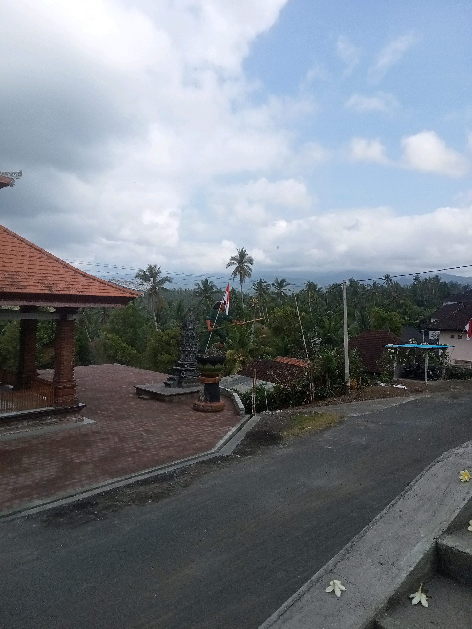A person stands on a raised platform in Mendoyo, Indonesia, holding a red and white Indonesian flag on a pole. To the left, a building with an orange tiled roof and ornate carvings stands on a brick-paved area. Behind the building and the flag-bearer is a lush green landscape of palm trees and other vegetation, with mountains visible in the distance under a cloudy sky. A paved road runs in front of the platform, with a concrete barrier along the edge. A few white flowers are scattered on the concrete.