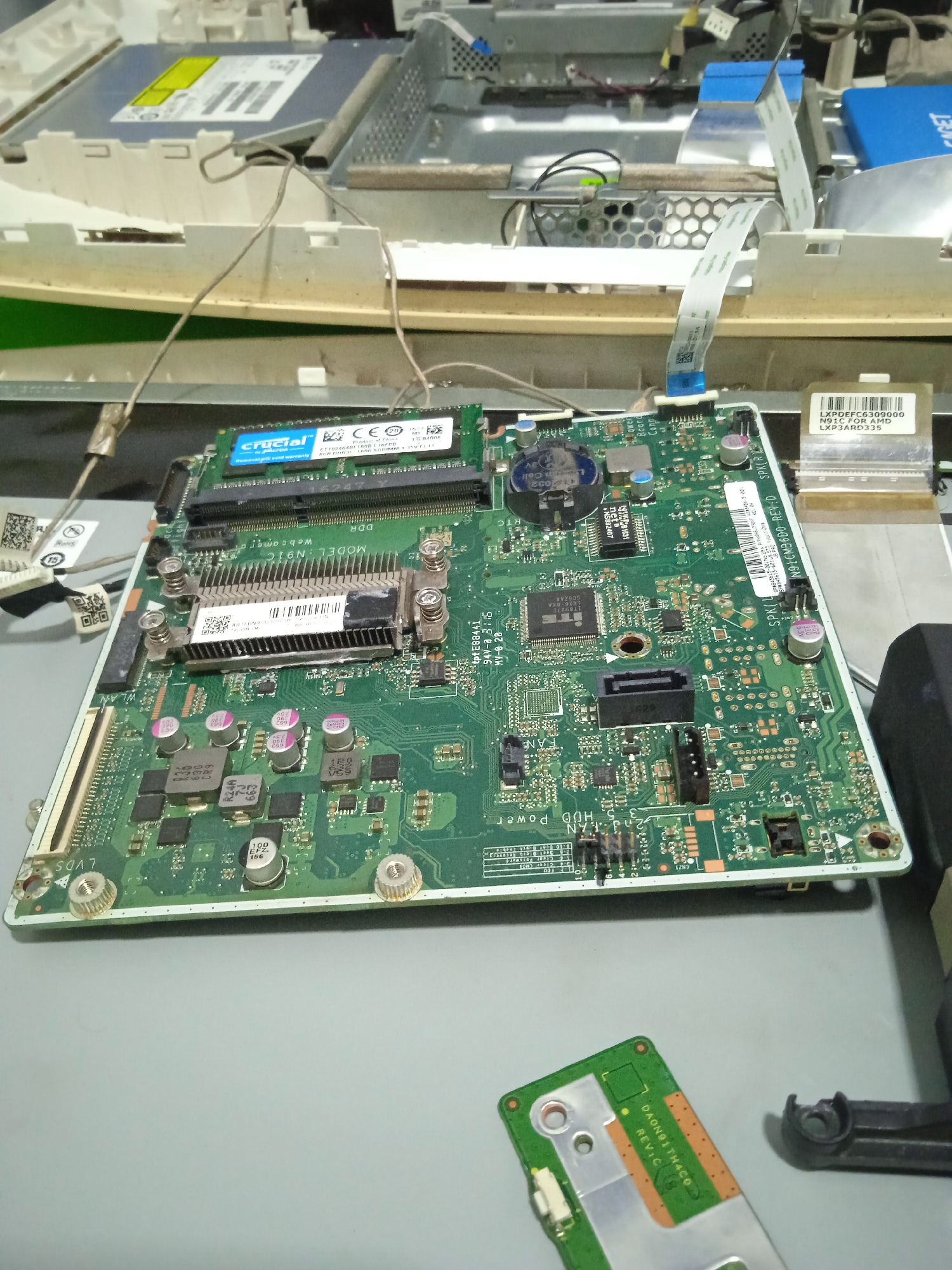 The image shows a disassembled computer motherboard lying on a gray surface. The motherboard is green and covered with various electronic components, including capacitors, resistors, and integrated circuits. A RAM stick is inserted into a slot on the motherboard. In the background, parts of the computer casing and other electronic components are visible. The scene appears to be a workspace or repair area for electronics. The image is taken from a slightly elevated angle, looking down at the motherboard. There are no people or discernible environmental features that would suggest a specific city or location. The image is devoid of any information to place it in Cileunyi, Indonesia.