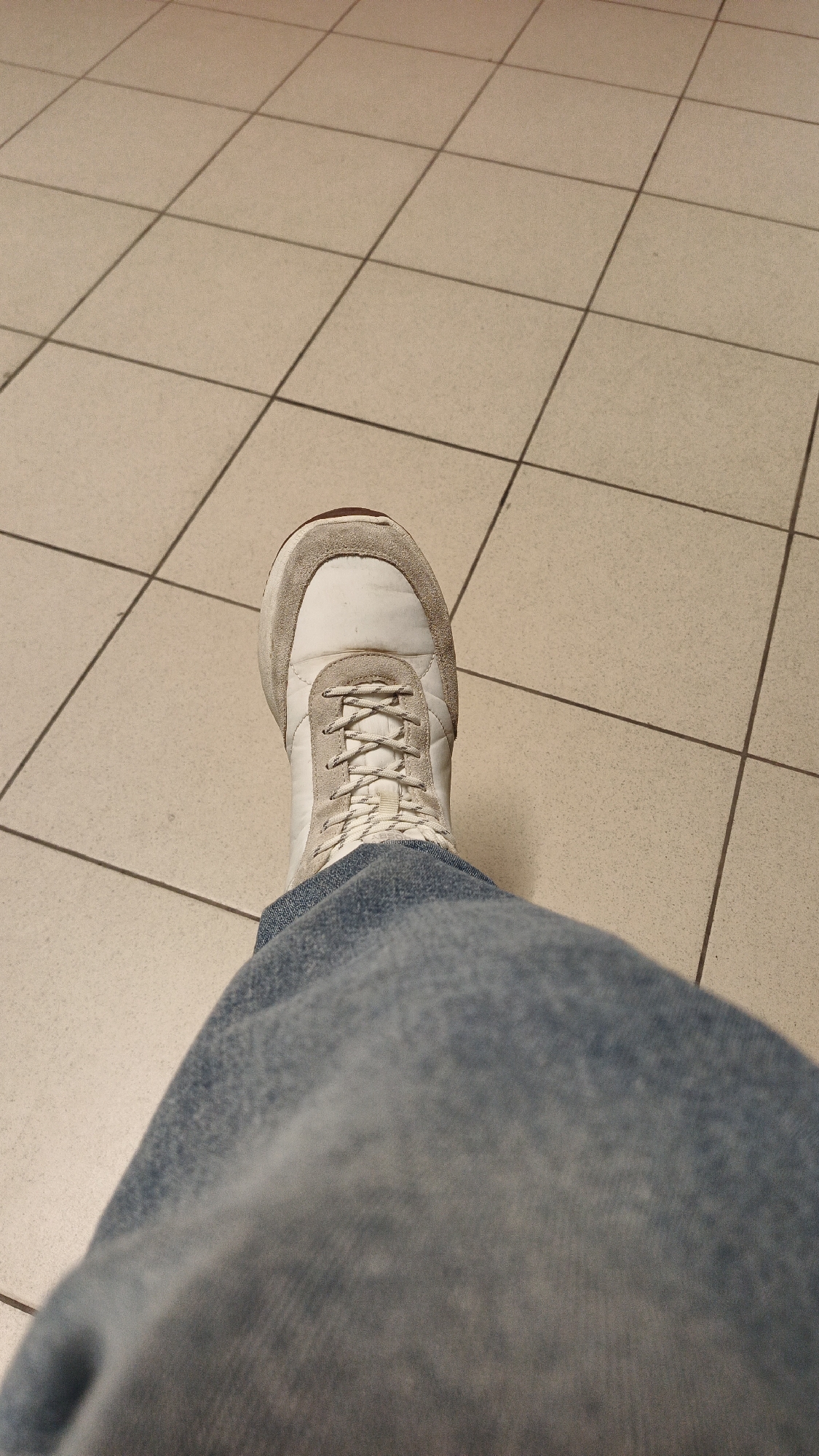 An extreme close-up shot shows a white and gray sneaker with white laces, worn by an individual whose blue jeans fill the bottom half of the frame. The sneaker is positioned on a light beige tiled floor, with dark grout lines creating a grid pattern. The lighting suggests an indoor setting, possibly a public space. The image does not provide any identifiable landmarks or context to determine a specific location within Yekaterinburg, Russia.