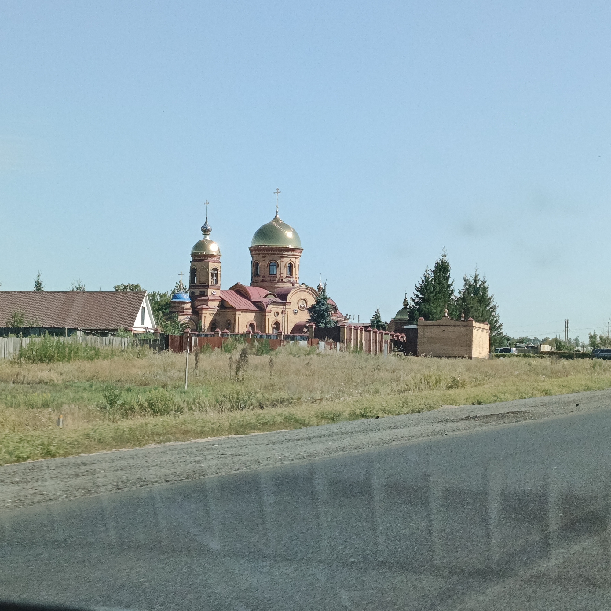 The image shows a rural landscape with a church and residential buildings. The church, made of brick with red roofs and golden domes, is the prominent structure. To its left, a house with a corrugated metal roof and a white wall is visible. A wooden fence runs along the front of the property. The foreground features a dirt or gravel road adjacent to a grassy field. Several trees, including tall evergreen trees, are scattered throughout the scene. The sky is clear and blue. The location is identified as Maiorskoe, Russia, implying a village or small town setting. There are no people or animals visible in the image. The overall impression is one of a quiet, possibly remote, rural settlement.