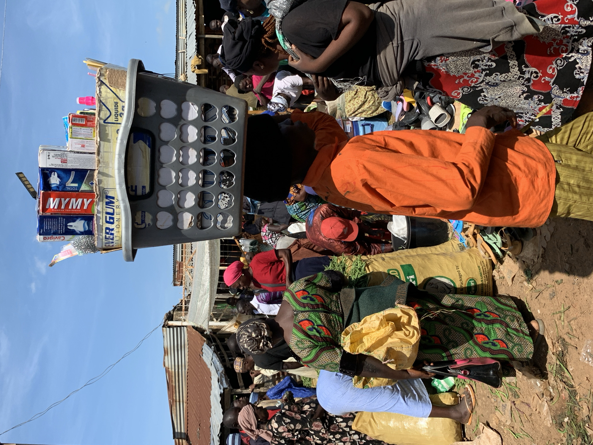 In Gura Top, Nigeria, a bustling outdoor market scene unfolds under a clear blue sky. A young person, dressed in an orange long-sleeved shirt and striped pants, carries a large grey plastic basket filled with various products on their head. The basket contains items such as boxes of toothpaste, soap, and other packaged goods. The individual is positioned in the foreground, facing slightly to the right, and appears to be interacting with another person.
Surrounding them is a diverse crowd of people, many of whom are women wearing colorful clothing and headscarves. Some individuals are engaged in transactions, while others are browsing goods. Large sacks, likely containing produce or grains, are visible on the ground, along with scattered debris. The market environment is characterized by makeshift stalls with corrugated metal roofs. The overall impression is one of vibrant commerce and daily life in a Nigerian community.