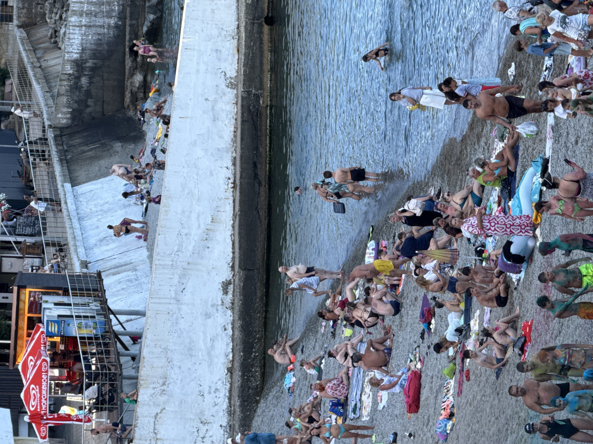 The image depicts a busy beach scene in Partenit, Ukraine, with numerous people enjoying the water and sand. The beach is pebbly, and the sea is calm. In the background, a concrete wall separates the beach from a higher level with a boardwalk, an ice cream stand, and what appears to be a cafe. People are seen sunbathing on towels, swimming in the sea, and walking along the shore. Some individuals are partially covered in brightly colored powder, suggesting a festive or recreational activity. The overall atmosphere is one of a summer day at a popular tourist destination.