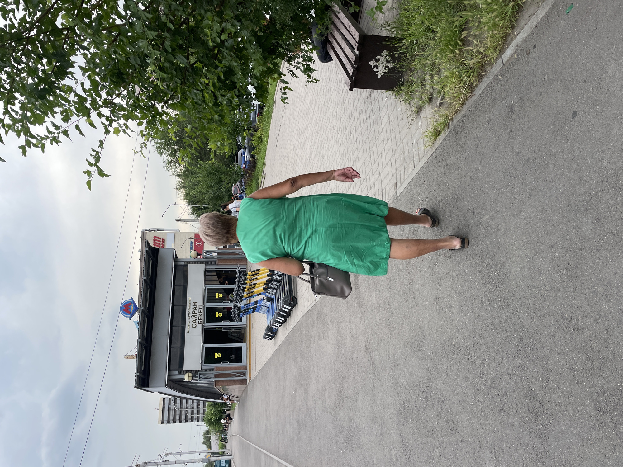 An elderly woman wearing a bright green dress and dark sandals walks away from the camera on a paved sidewalk. She carries a large gray handbag in her left hand. To her left, a metro station entrance is visible, with signs in Cyrillic script reading "Сайран Бекети" (Sairan Station). Several electric scooters are parked in front of the station entrance. The environment appears to be a city street with trees and buildings in the background. The sky is overcast. A wooden bench with an ornate design is situated to the right of the sidewalk, partially obscured by foliage.