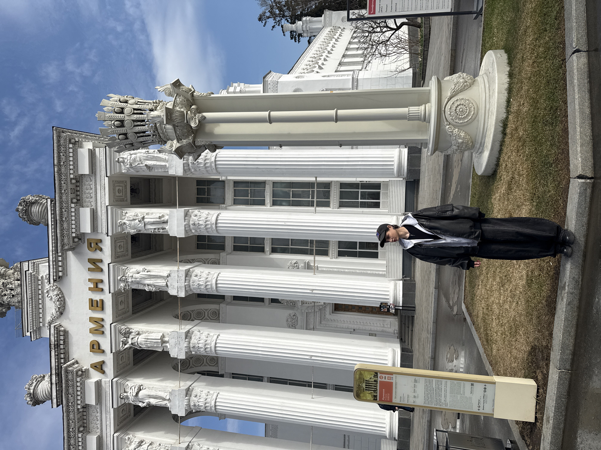 A person stands in front of the Armenia Pavilion at VDNH, Moscow, Russia. The pavilion is white with prominent columns and decorative sculptures. "Армения" is displayed in gold lettering above the entrance. An information sign is visible nearby. The sky is partly cloudy.