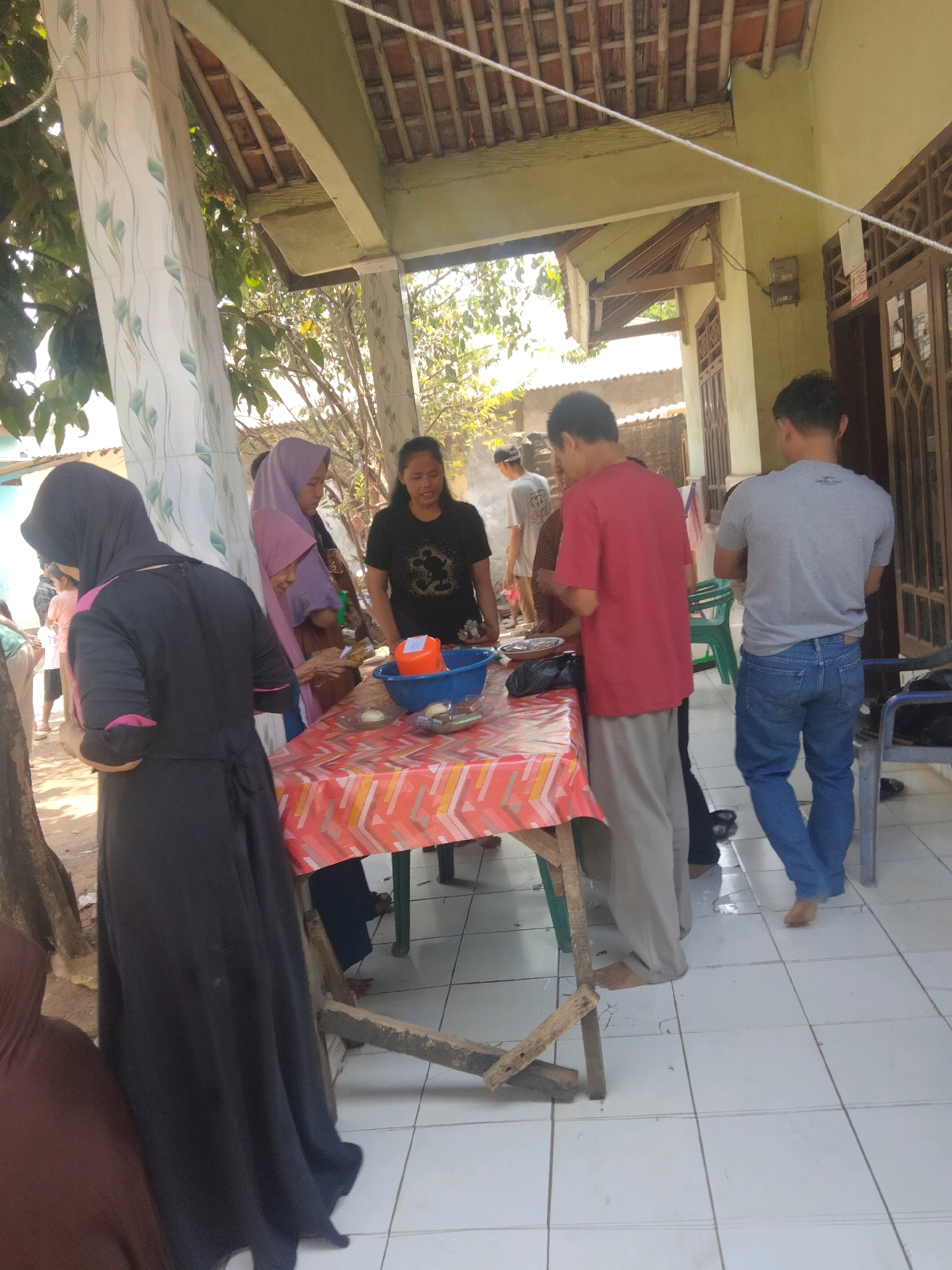In Suka Sari, Indonesia, a group of people are gathered outdoors under a covered patio with a tiled floor. The patio has a light green painted wall and a roof constructed from wooden beams and tiles. To the left, a patterned pillar supports the roof. A table with a colorful, patterned tablecloth is set up, laden with food items in bowls, including a blue bowl containing an orange container and other dishes.
Several individuals are interacting around the table. A woman wearing a dark hijab and a black dress with pink trim stands at the left, facing away from the camera. Next to her, a woman in a purple hijab and a brown patterned top is looking at something on the table. To her right, a woman in a black t-shirt with a Mickey Mouse graphic is talking.
Further back, a man in a red shirt and light gray pants stands behind the table. To his right, another man in a gray t-shirt and blue jeans is standing. Other individuals are visible in the background, some partially obscured. The environment suggests a social gathering, possibly a meal or a communal event. The ground around the patio appears to be dirt.