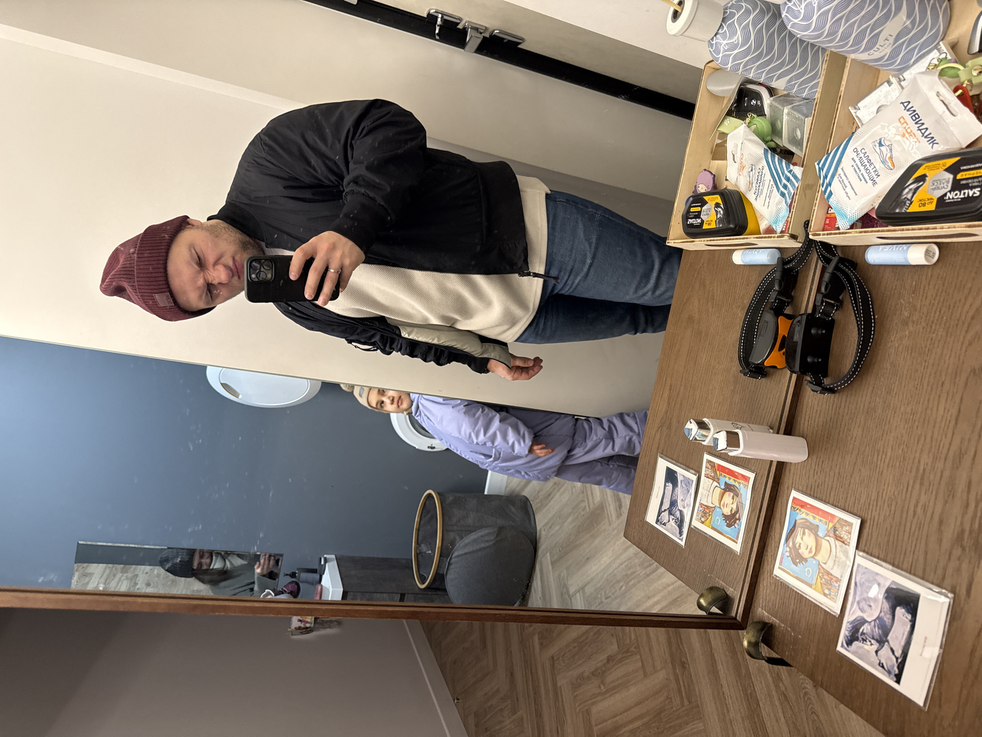 A man wearing a burgundy beanie, black jacket, white hoodie, and blue jeans is taking a selfie in a mirror. Reflected in the mirror is a woman wearing a light purple jumpsuit. The scene is indoors, likely in a home or apartment.
To the right of the mirror, a wooden surface is cluttered with various items including two wooden boxes containing cleaning products and supplies, lip balm, a dog collar, a white cylindrical object, and several small photographs.
In the mirror's reflection, behind the woman, there's a grey laundry hamper, a washing machine, and a blue wall.