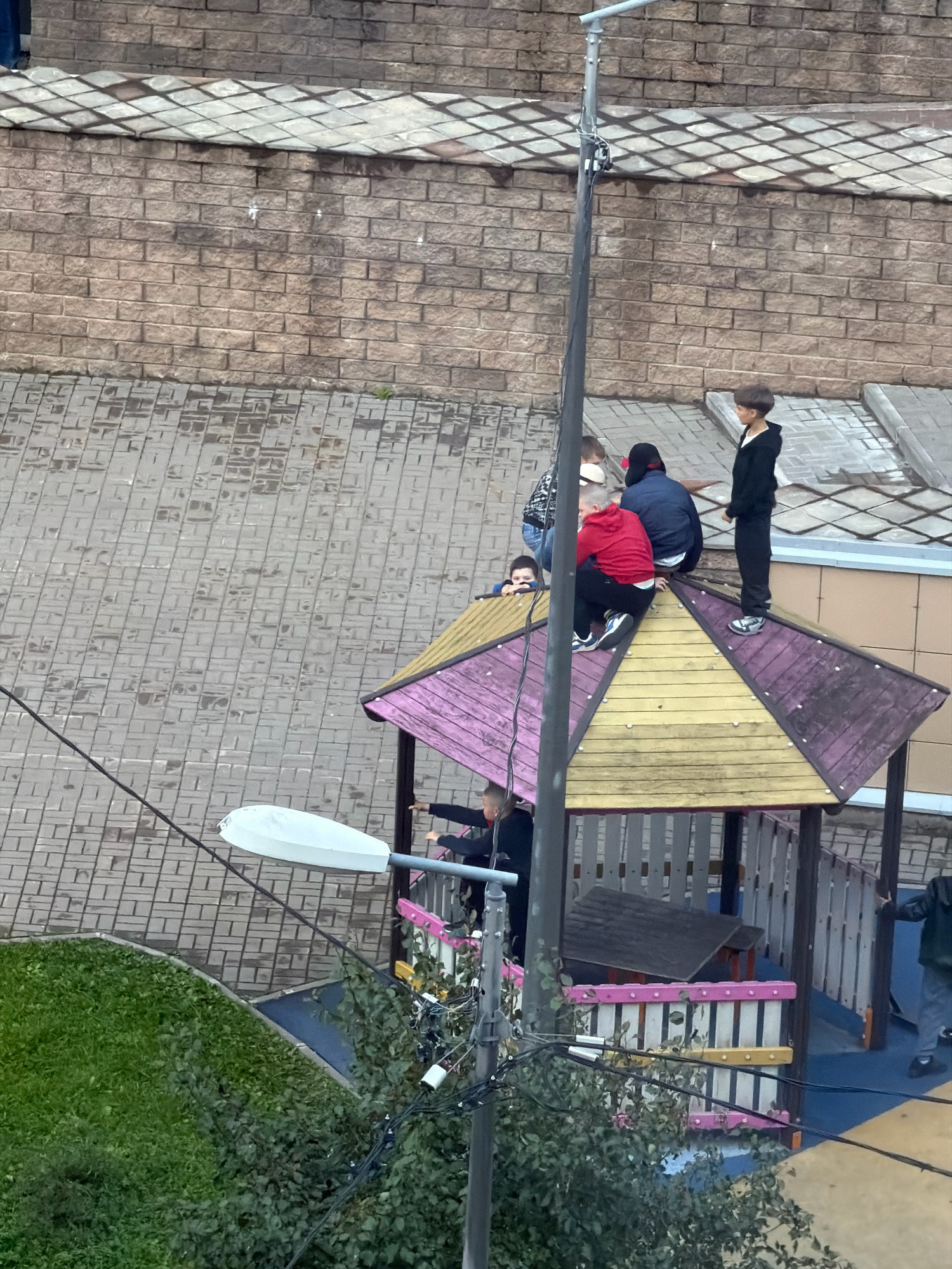 This image depicts an outdoor scene with a group of children on and around a colorful playground structure. The playground has a peaked roof with alternating purple and yellow sections. Several children are visible on the roof, including one in a black hoodie and tracksuit standing on the right, and another in a red jacket and black pants on the left. A street lamp is positioned in the foreground, with its pole running vertically through the image. In the background, there is a brick wall and a tiled roof. To the left of the playground is a patch of green grass and dense green foliage. The ground is paved with gray rectangular tiles. The overall impression is a daytime scene in an urban or suburban environment. The city of Unknown, Russia is specified for the location.