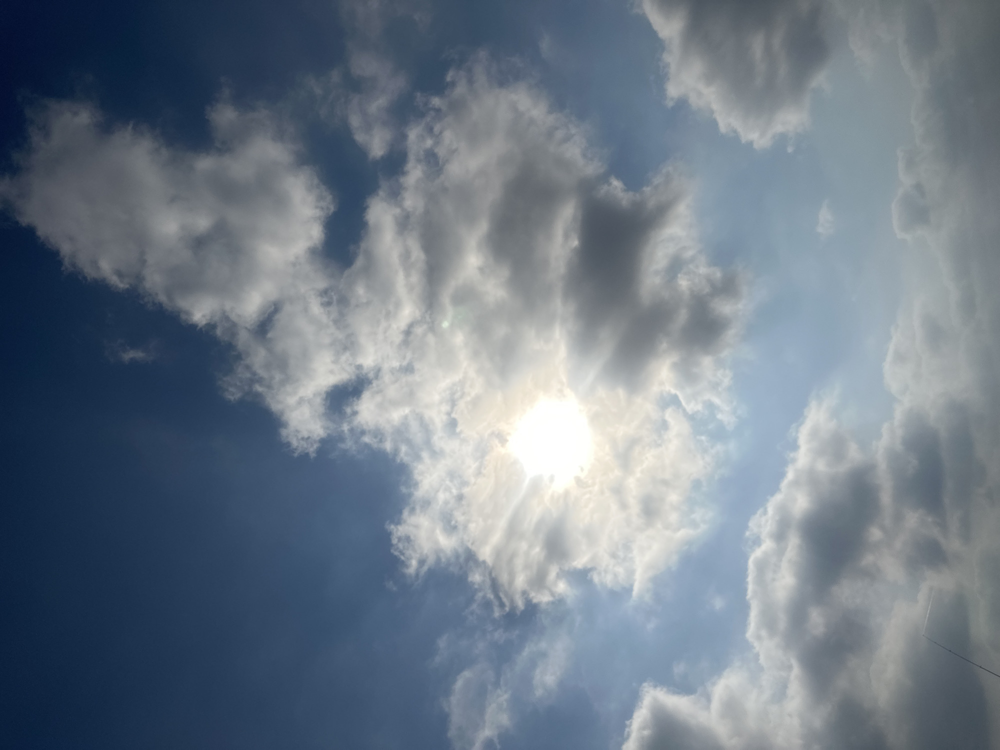 The image captures a bright, sunny sky over Haifa, Israel, characterized by a mix of fluffy white and darker gray clouds. The sun is prominently featured, partially obscured by clouds, casting rays of light that illuminate the sky and create a luminous effect. The clouds are varied in size and density, with some appearing wispy and others more substantial and shadowed. The overall impression is of a dynamic sky with good visibility, typical of a clear day with some cloud cover in the region.