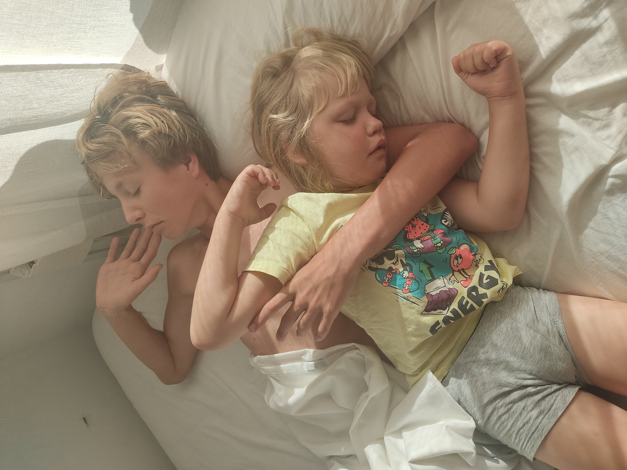 Two young children, a boy and a younger child, are sleeping in a bed. The boy, positioned on top, is lying on his back with his right arm bent and his hand covering his mouth. His eyes are closed, and his hair is blond and messy. The younger child, lying below the boy, is on their left side, facing away from the viewer, with their eyes closed and blond hair visible. This child is wearing a yellow t-shirt with graphics and grey underwear. The bed is covered in white sheets, and sunlight streams in from the right, casting shadows from a curtain onto the scene. The overall setting appears to be a bedroom in a domestic environment, possibly in the city of Blanes, Spain, given the context.