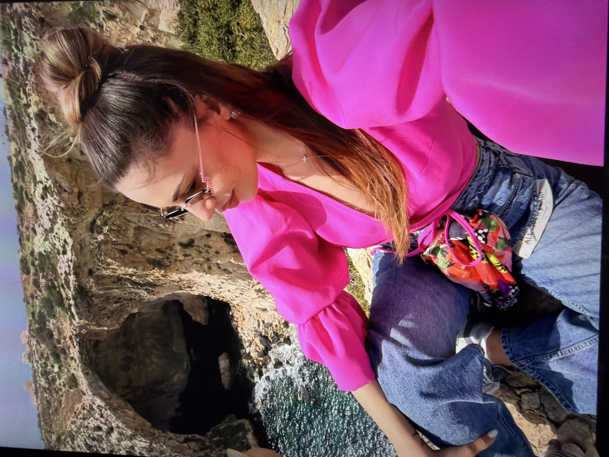 An eye-level shot captures a young woman outdoors on a bright day, positioned on what appears to be a rocky coastline overlooking the sea. She is seen from the chest up, leaning forward slightly and looking down. Her long brown hair is styled in a messy bun, and she wears sunglasses with pink frames.
She is dressed in a vibrant fuchsia or hot pink blouse featuring voluminous, puffed sleeves and a V-neckline. A delicate silver cross necklace is visible around her neck. She is also wearing blue denim jeans, and tucked into her left hip is a colorful, patterned accessory, possibly a small bag or scarf, with red, black, and white motifs.
The background reveals rugged rocky terrain, with a prominent dark cave opening visible below and to the left of the woman. Turbulent blue-green ocean water, showing whitecaps, can be seen at the base of the rocks. The overall setting suggests a scenic coastal location with natural rock formations and sea caves. The lighting indicates it is daytime, likely sunny. On a white tag or fabric piece attached to her jeans, faint text reads "ALL-OV" and "OVER AGAIN." The woman appears relaxed, enjoying the expansive view.