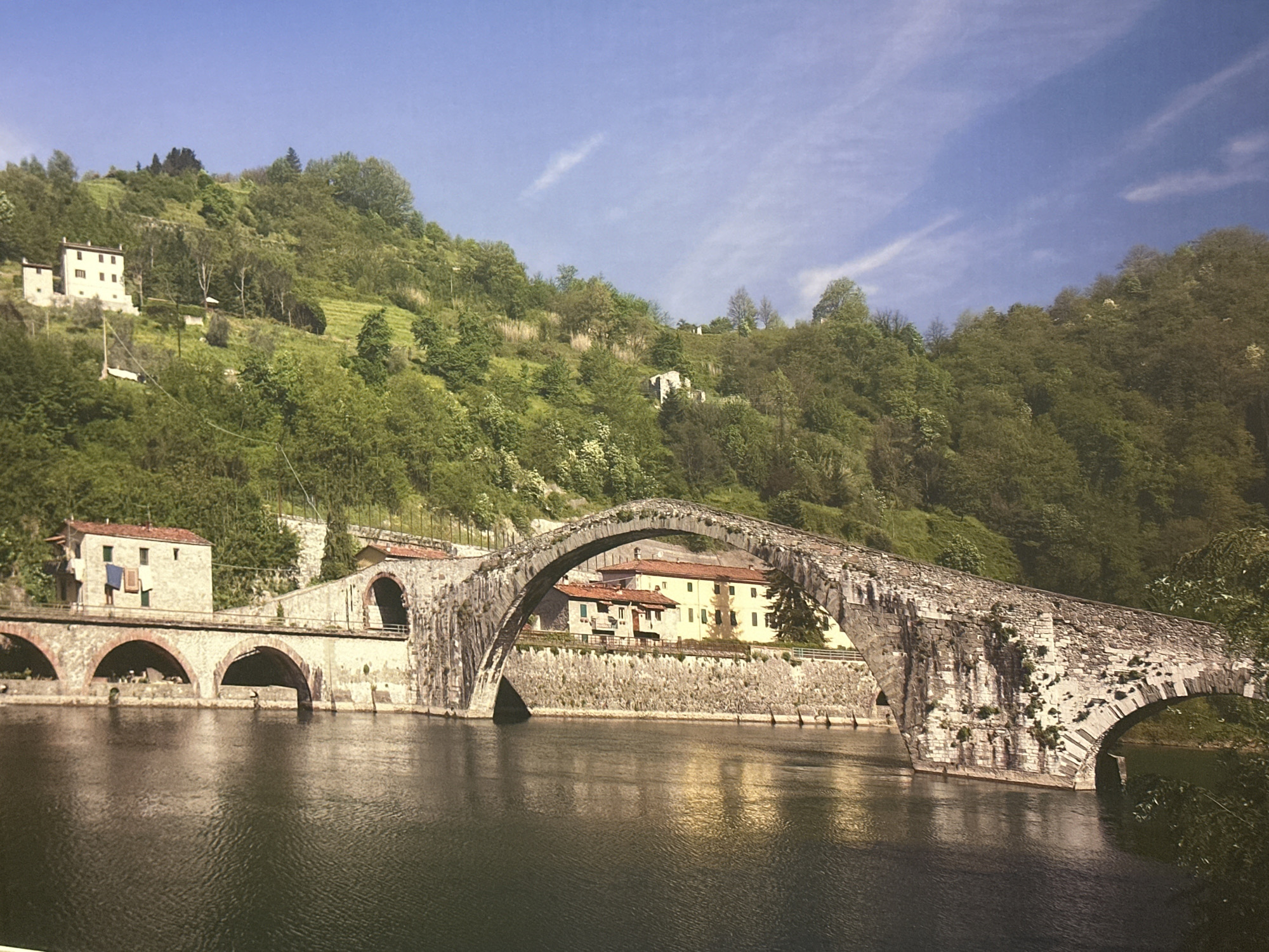 The image depicts a stone bridge with multiple arches spanning a body of water. The bridge is made of weathered stone and appears to be ancient. On the far bank of the water, there are buildings with red tiled roofs nestled into a lush green hillside. Trees and vegetation cover the slopes of the hill, and the sky is a clear blue with a few wispy clouds. The water reflects the bridge and the surrounding landscape. This scene is not located in Harrisburg, United States of America.